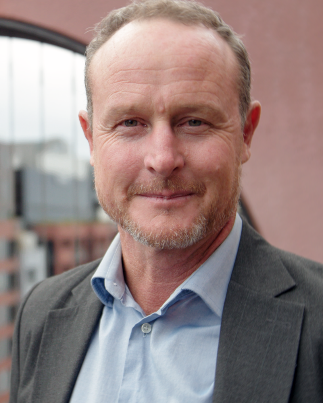 A middle-aged man with a trimmed beard and light brown hair, smiling, wearing a light blue shirt and a dark suit jacket, standing outdoors with a building in the background.