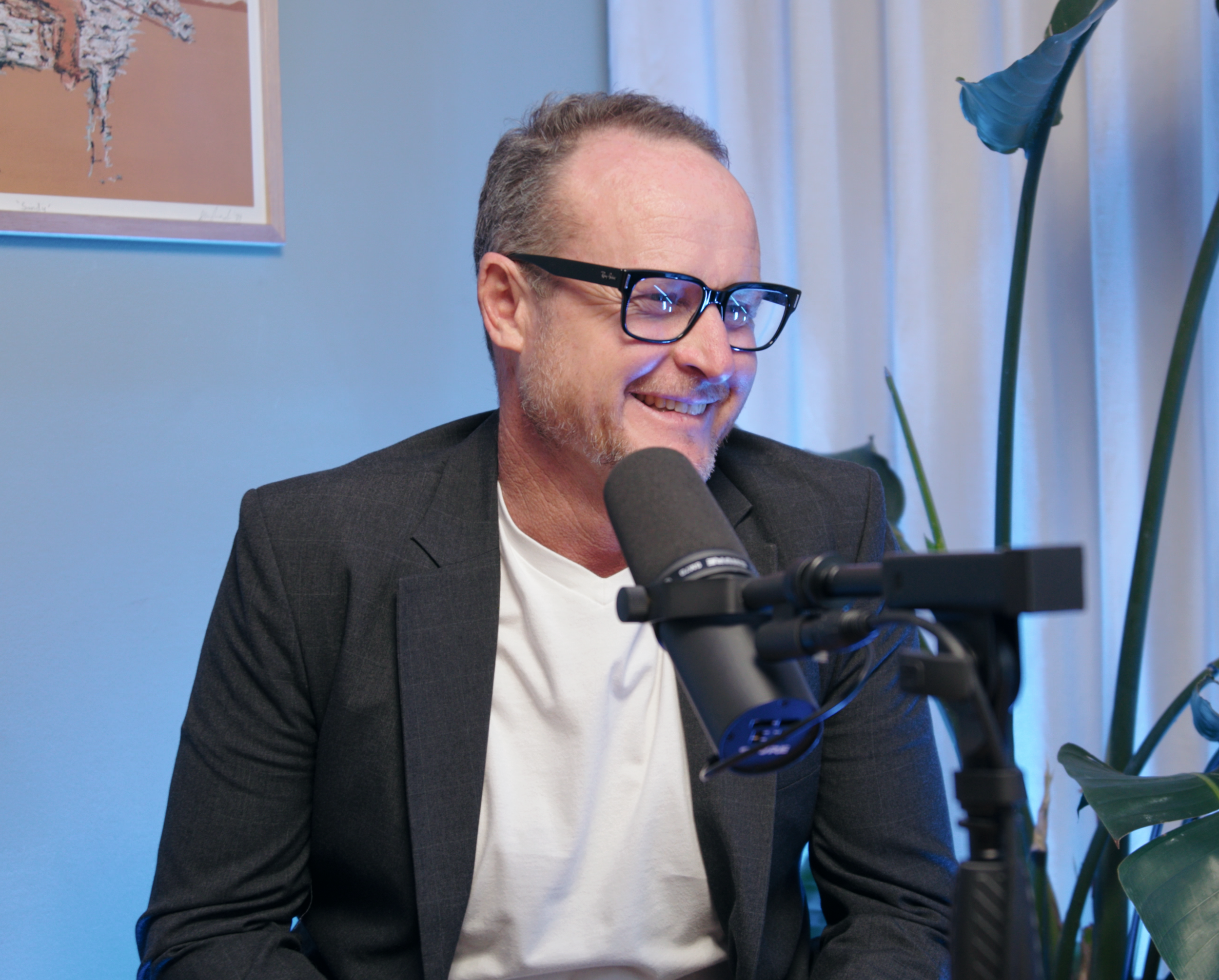 Smiling man with glasses speaking into a microphone during a recording or interview in a room with artwork on the wall and large green plants.
