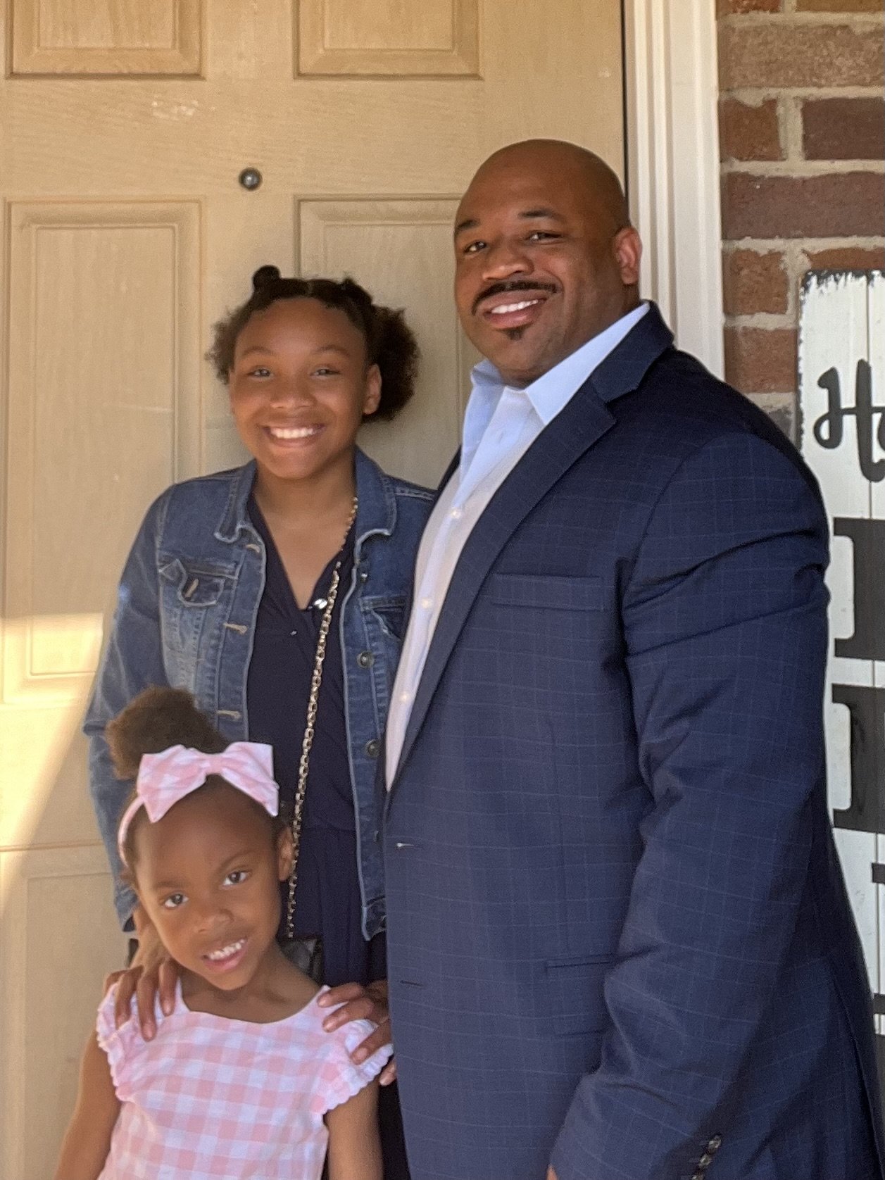 A man in a suit standing with two young girls in a home setting, all smiling at the camera.