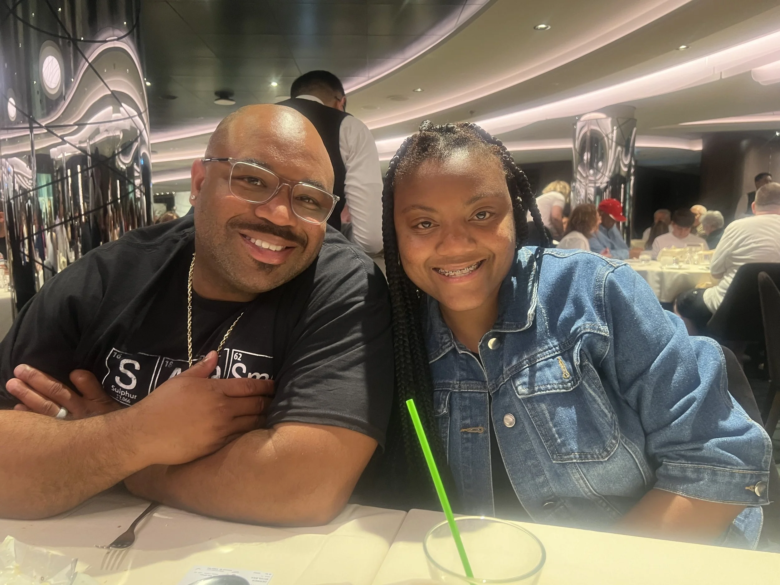 A man and a woman sitting at a restaurant table, smiling at the camera. The man has glasses and is wearing a black T-shirt with jewelry, and the woman has braided hair and is wearing a denim jacket.