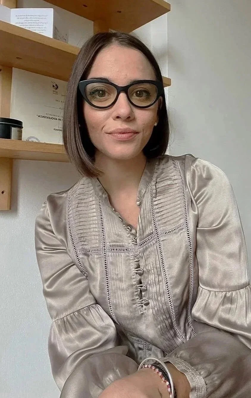 Una donna con capelli corti, indossa occhiali spessi e un vestito di seta beige, seduta vicino a una parete con scaffali di legno.