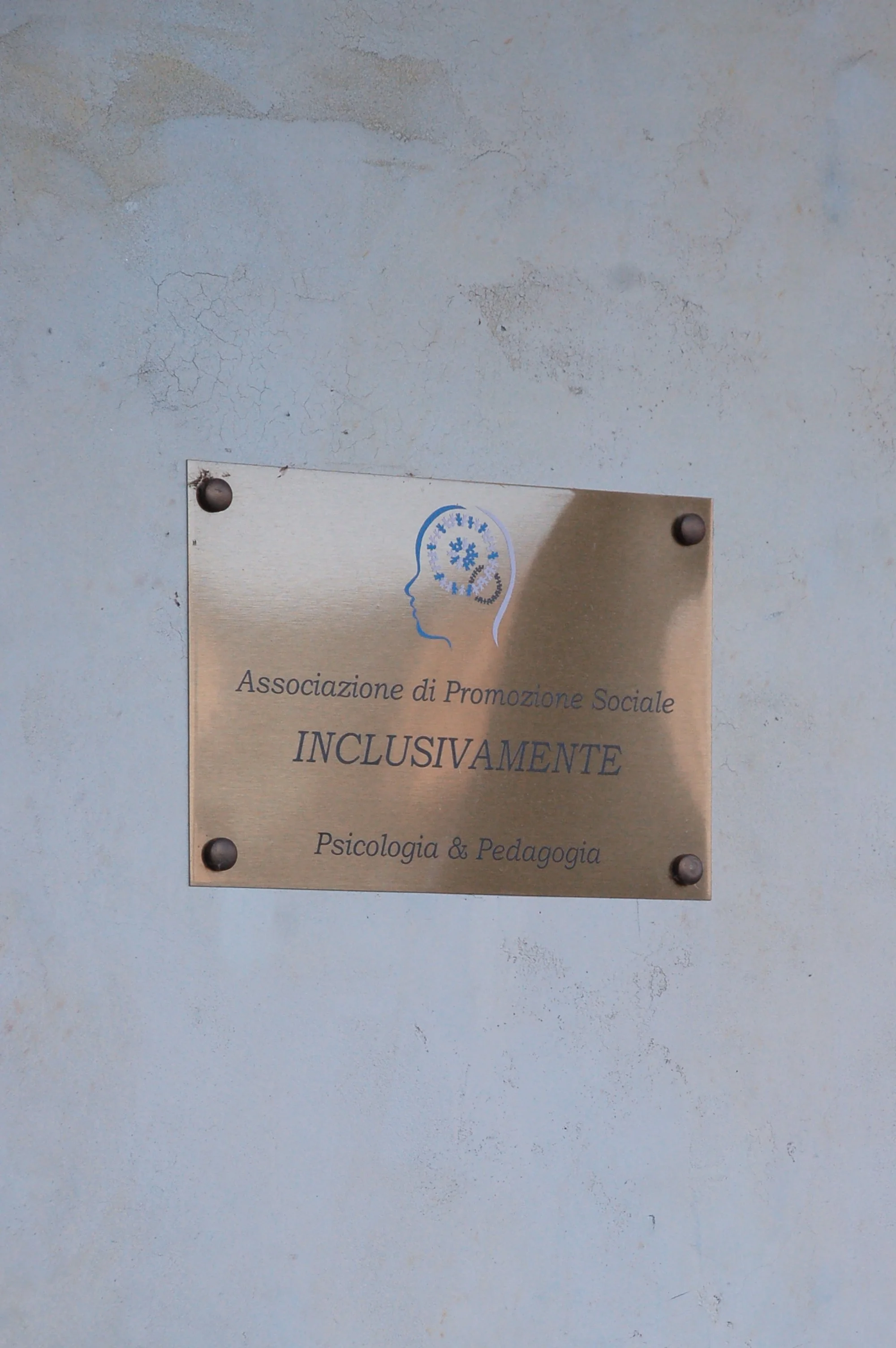Piastra in metallo con logo di una testa umana e testo in italiano su una parete di colore grigio con segni di usura.