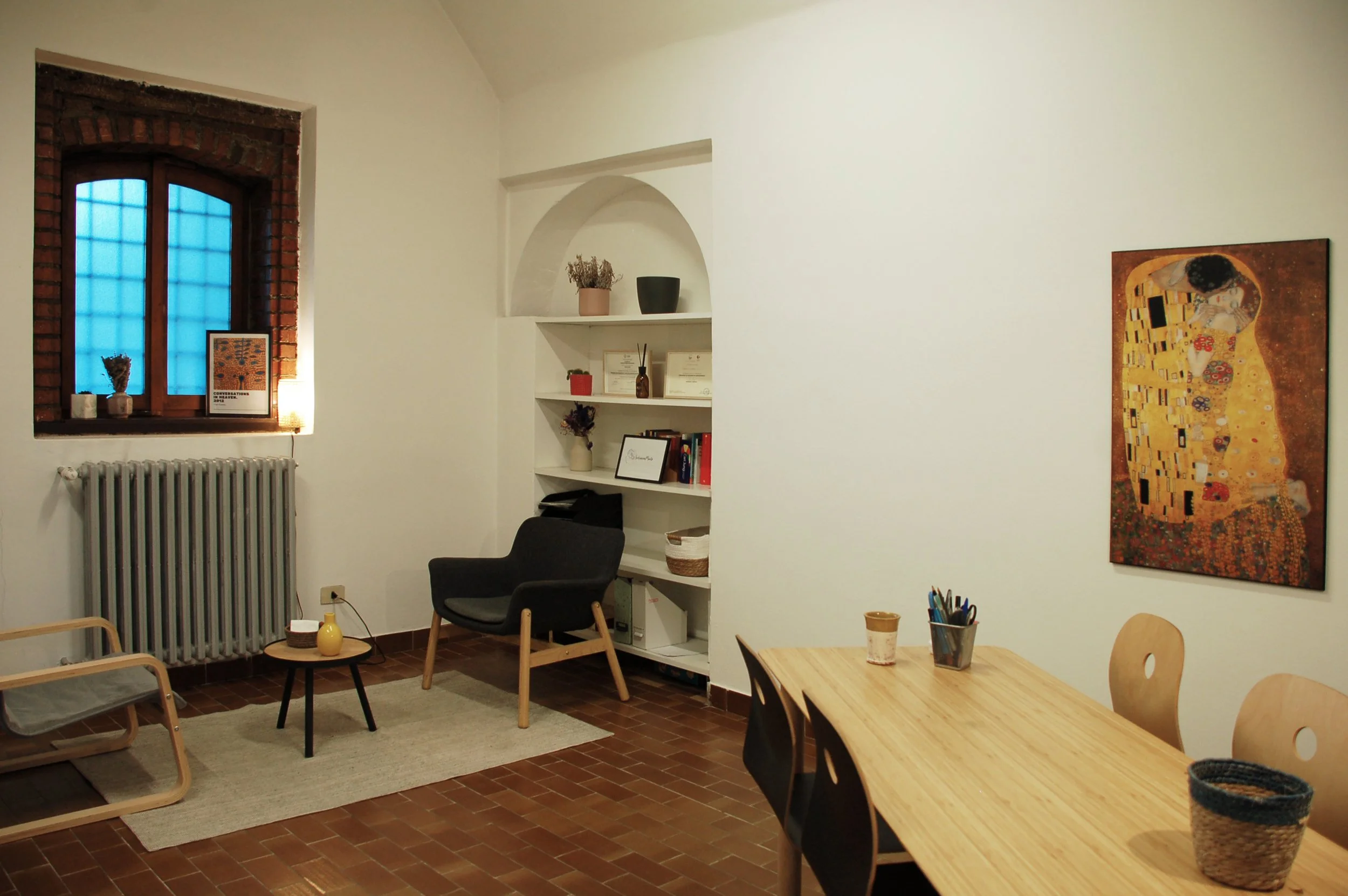 Interno di una stanza con pareti bianche, una finestra con cornice in mattoni, un divano nero, una sedia di legno e una sedia di plastica, un tavolo di legno con ciotole e penne, scaffali con libri e decorazioni, una casa con la famosa stampa di Klim