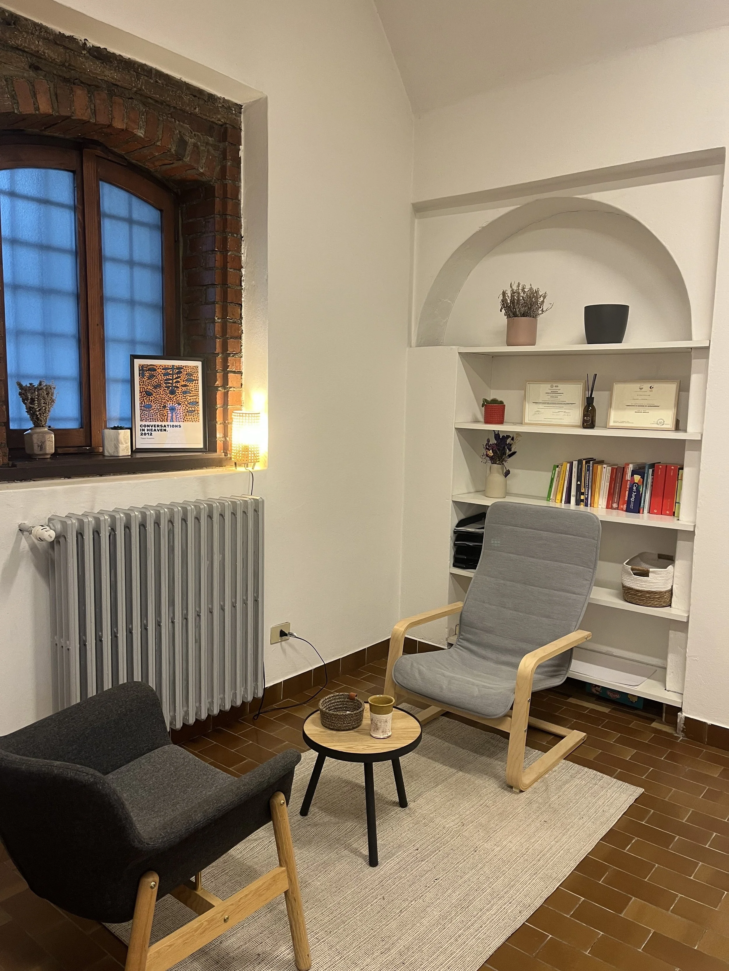 Angolo di soggiorno con sedia in legno e tessuto grigio, poltrona nera, tavolino rotondo in legno con oggetti decorativi, libreria bianca con libri, piante e quadri, finestra con cornice in mattoni e tende blu, tappeto beige sulla piastrelle marroni.