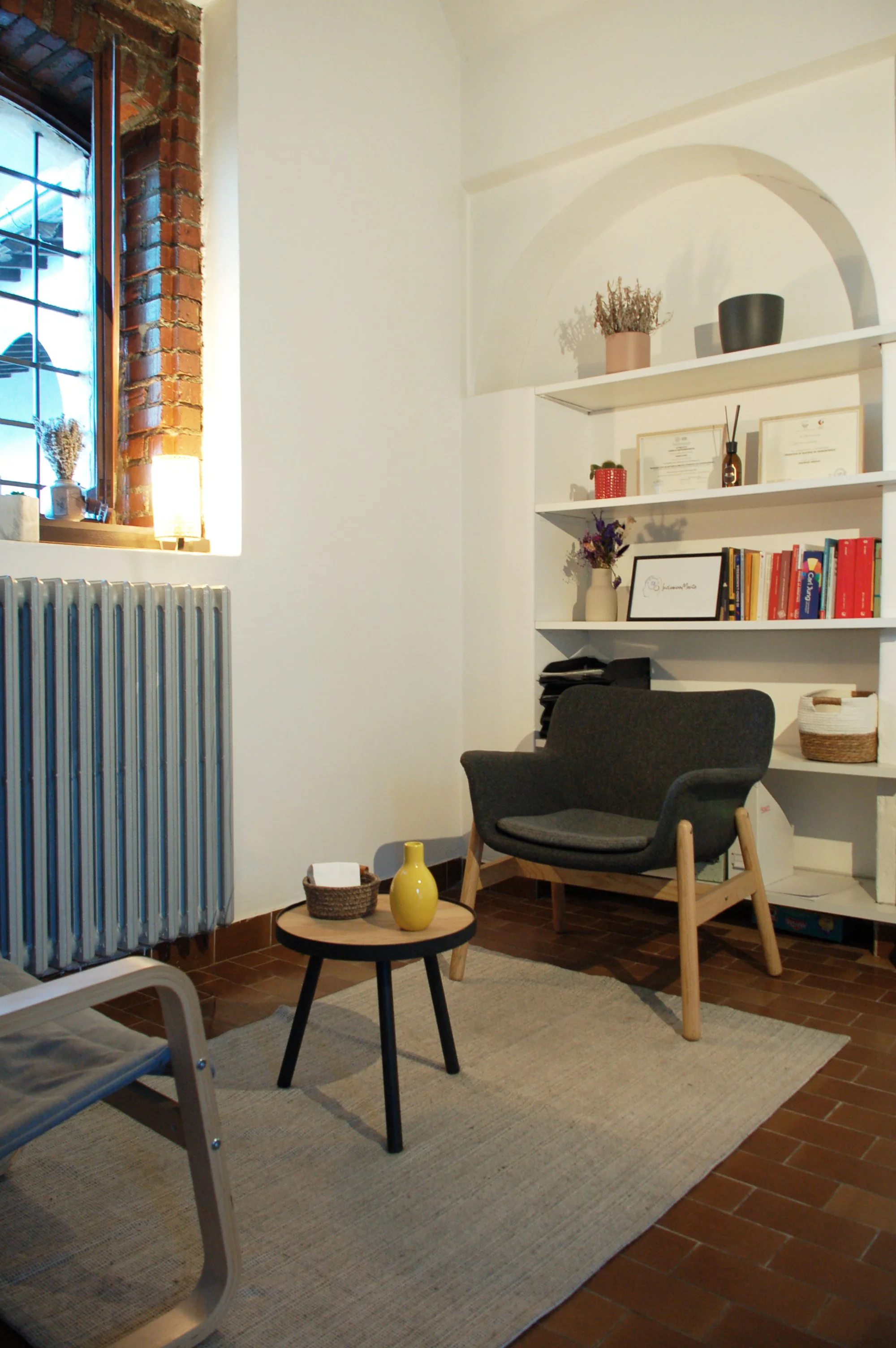 Soggiorno con sedia, tavolino, librerie, oggetti decorativi e finestra con piantine.