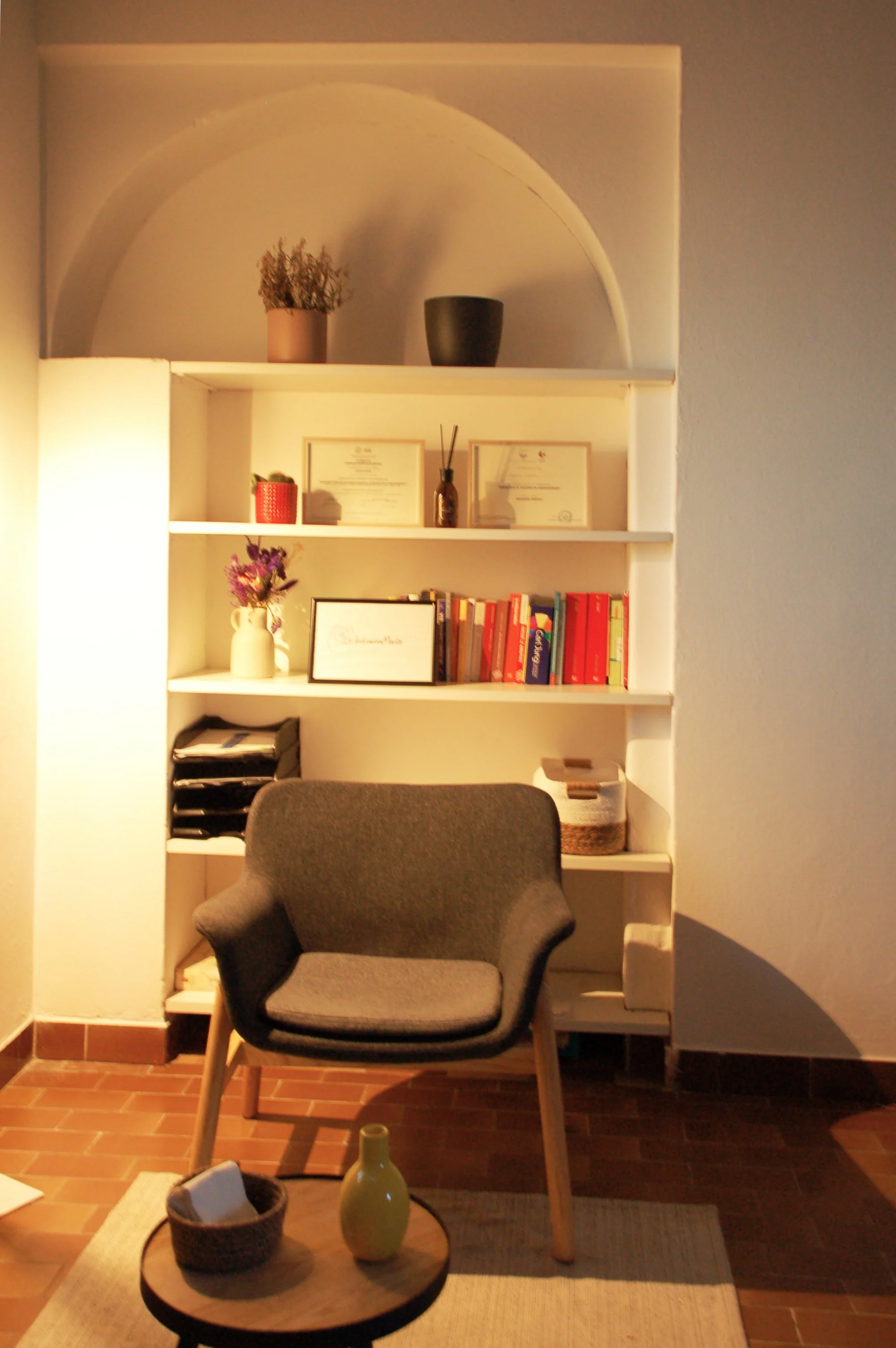 Soggiorno con sedia moderna grigia, libreria bianca con libri, vasi e decorazioni, pareti bianche con nicchie architettoniche, pavimento in piastrelle di ceramica marroni, tazzina e vaso giallo chiaro sul tavolino vicino alla sedia.