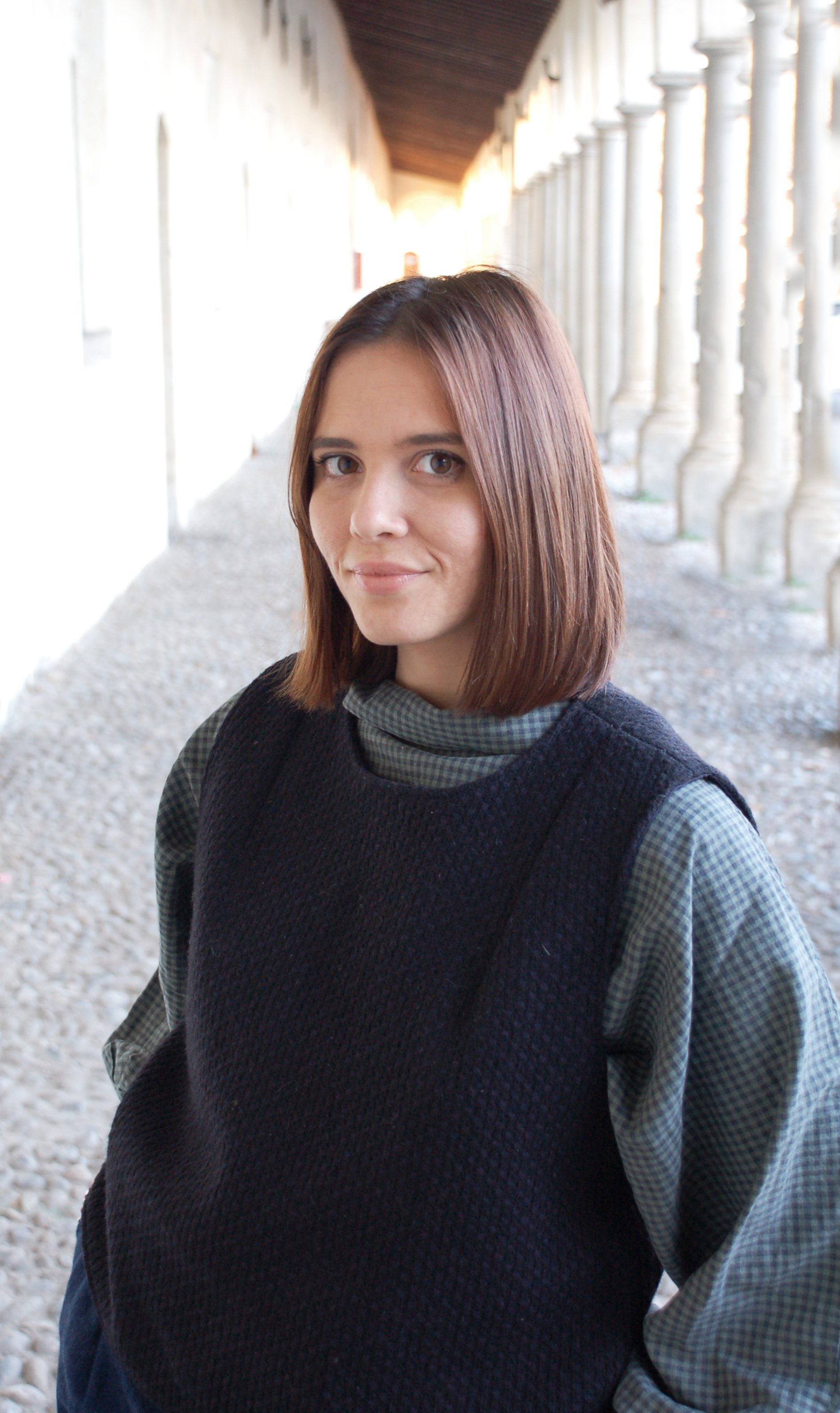 Ragazza con capelli corti castani, indossa una maglia a quadri e un maglione nero, si trova sotto un portico con colonne.