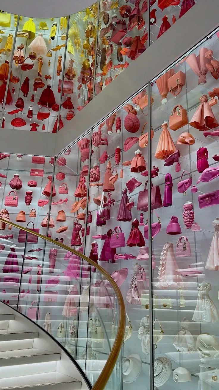 Colorful display of handbags, shoes, and accessories arranged on glass shelves inside a modern store with a staircase and glass walls.