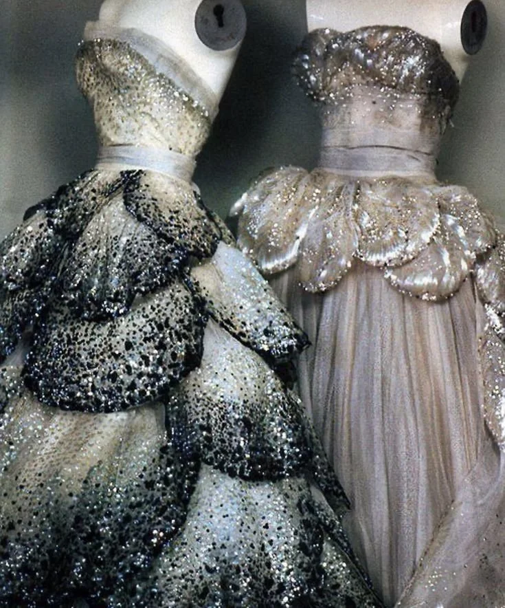 Two mannequins dressed in glamorous, sparkly gowns, one in a silver, black-studded dress and the other in a shimmering pearl-colored gown with ruffled details.