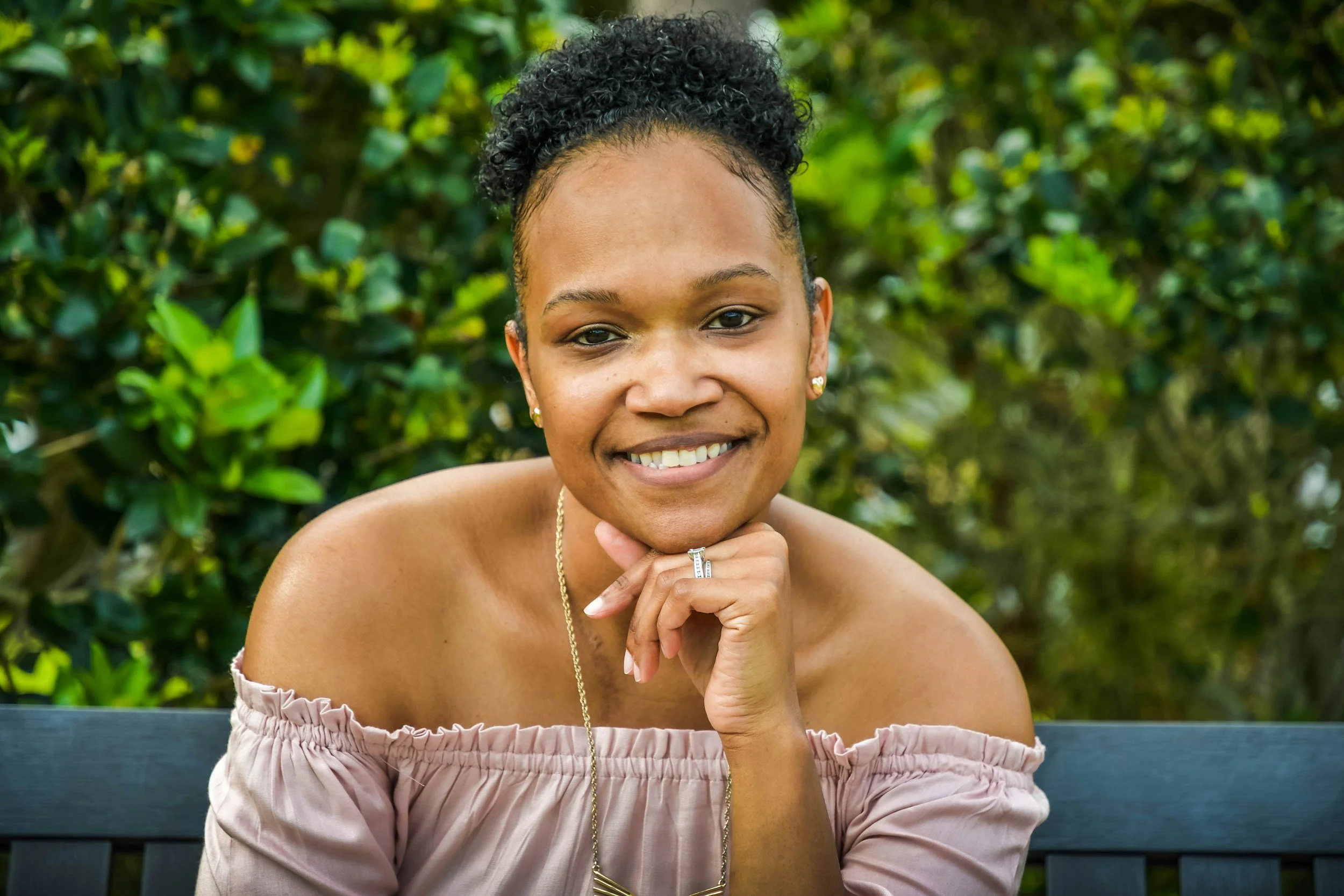 A smiling woman with short curly hair, wearing a pink off-the-shoulder top, earrings, and a necklace, sitting on a bench with greenery in the background.