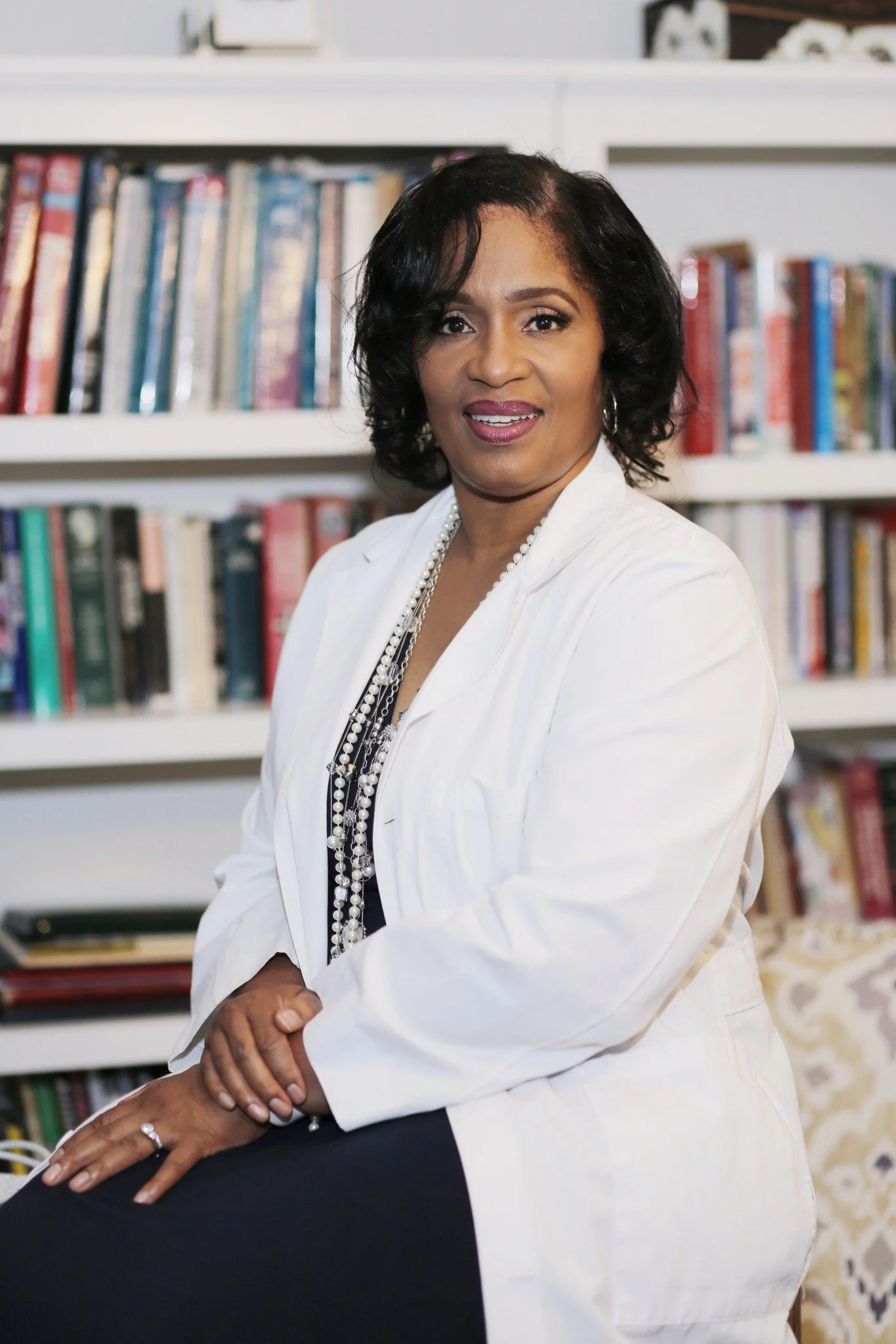 A woman with black, shoulder-length hair, wearing a white blazer and a long pearl necklace, sitting in a room with a bookshelf filled with books behind her.