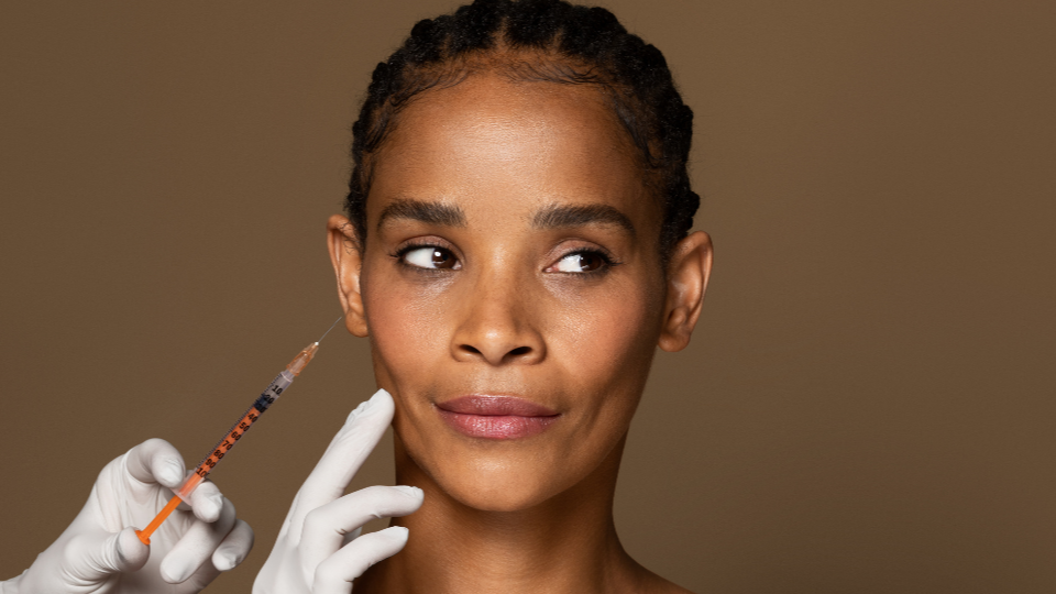 A woman receiving a cosmetic injection in her cheek from a medical professional wearing gloves.