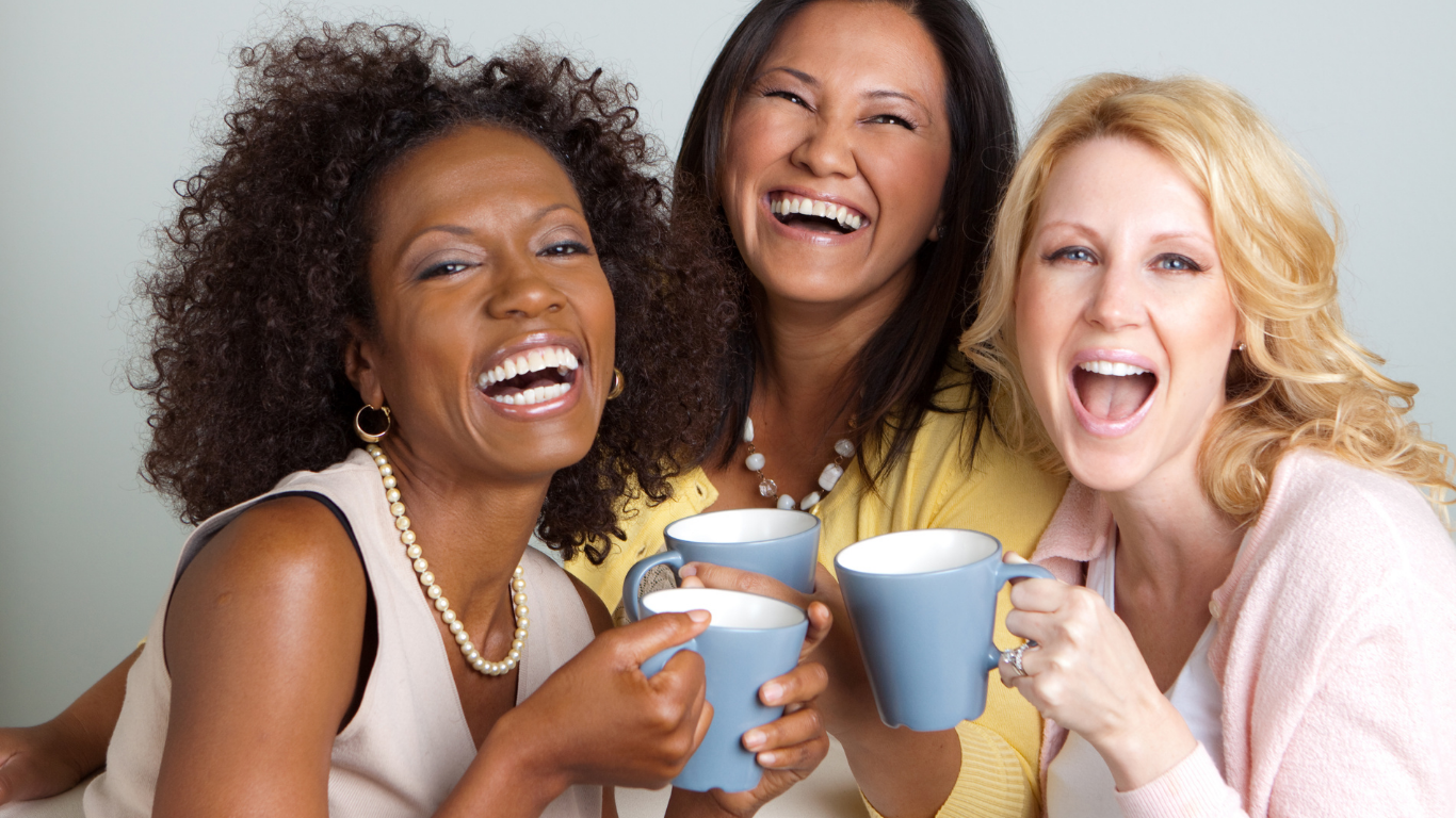 Three women smiling and laughing, holding blue coffee mugs, enjoying a happy moment indoors.