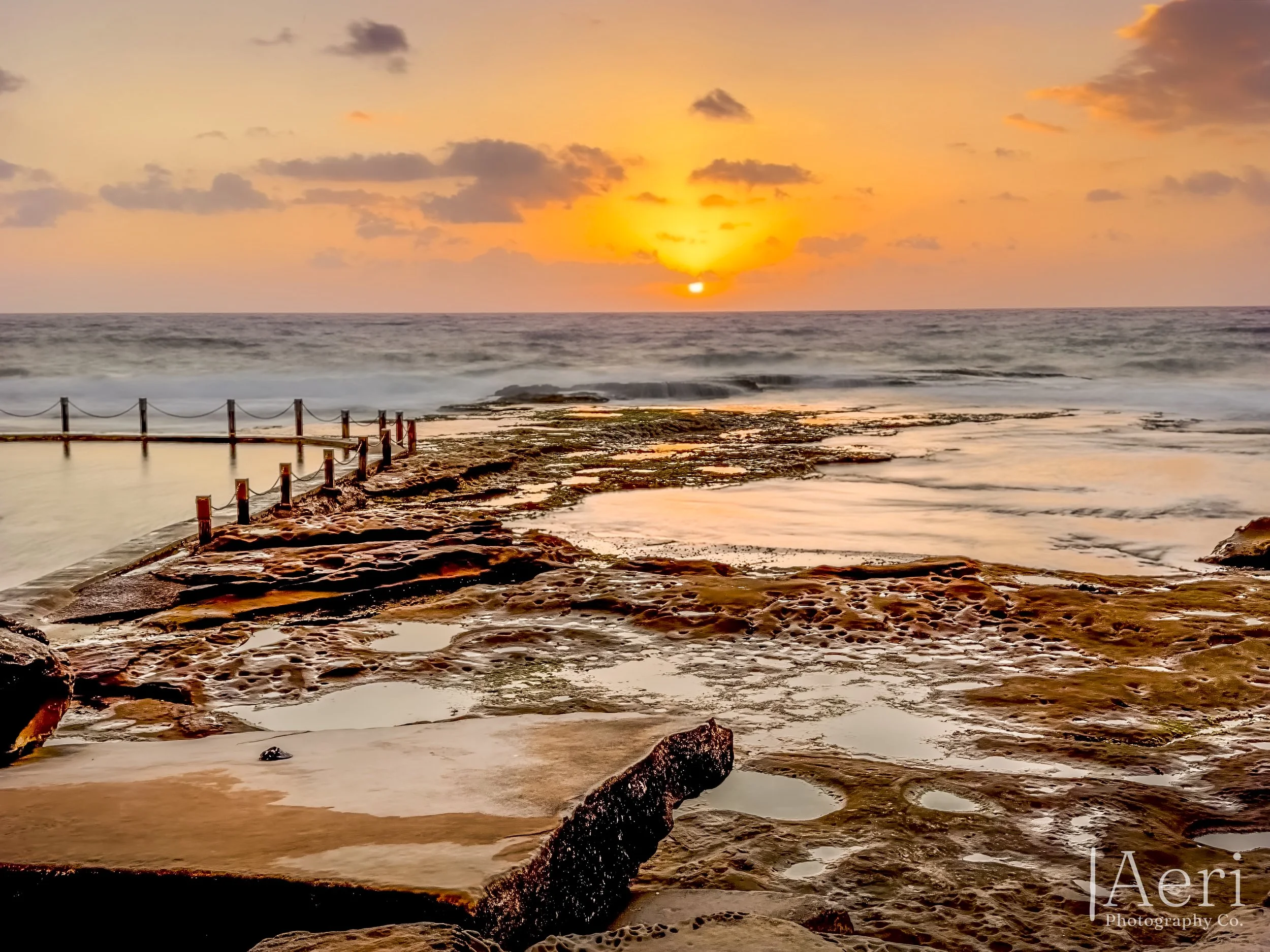 Sunset over the ocean with a rocky shoreline and tide pools in the foreground.