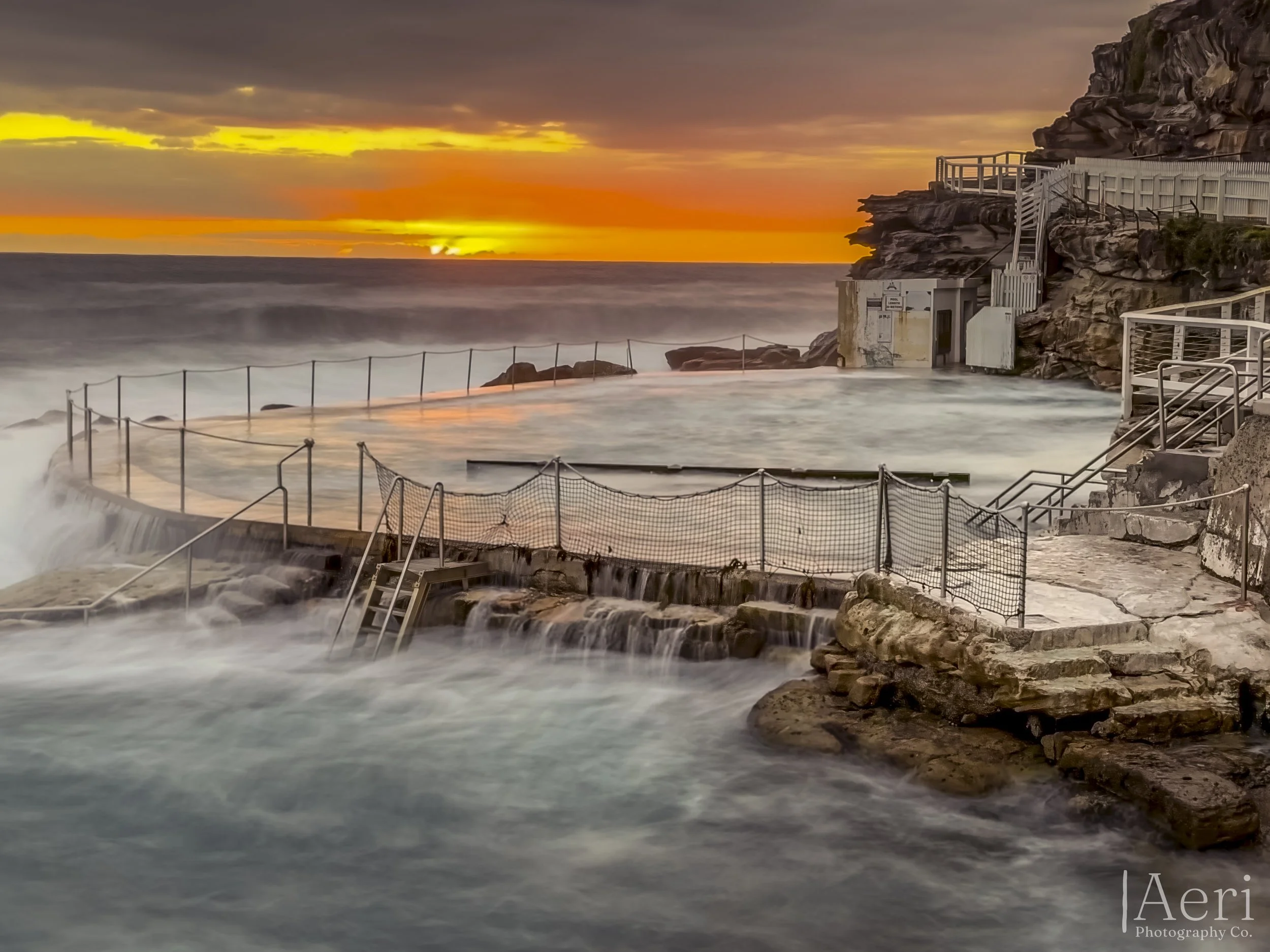 A coastal ocean pool with surrounding steps, railings, and a small building at sunset, with the ocean and sky in the background.