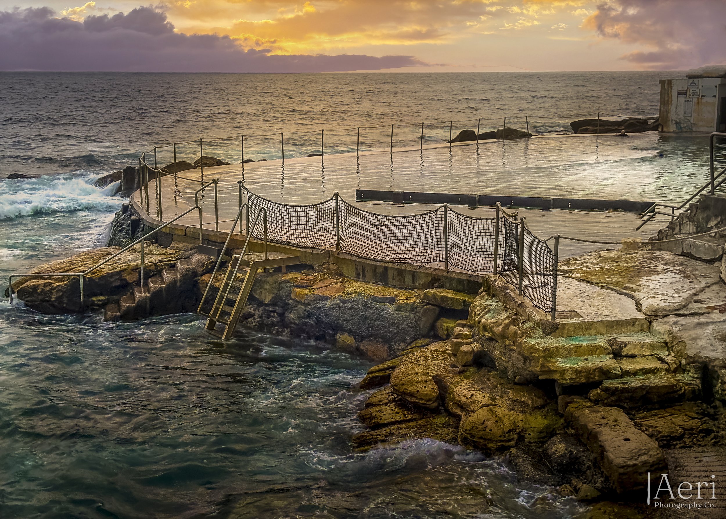 The image shows an outdoor hot spring pool with metal railings, built on a rocky shoreline, overlooking the ocean during sunset with a sky filled with clouds and warm colors.