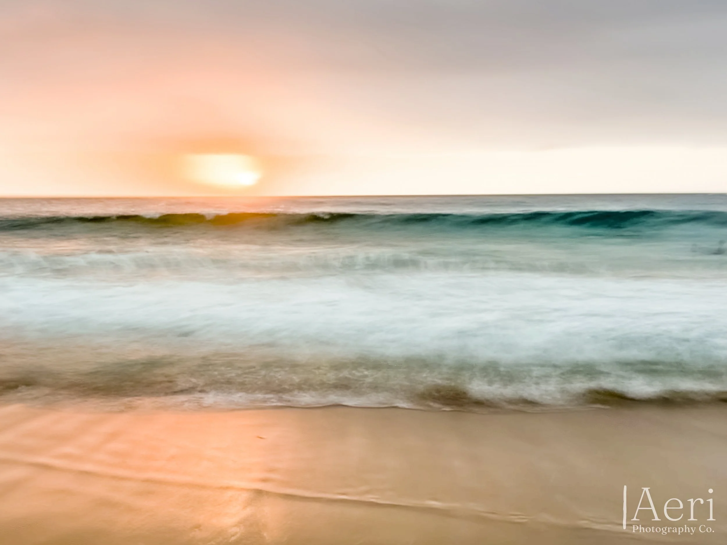 Sunset over the ocean with calm waves crashing onto a sandy beach, soft pastel sky colors.