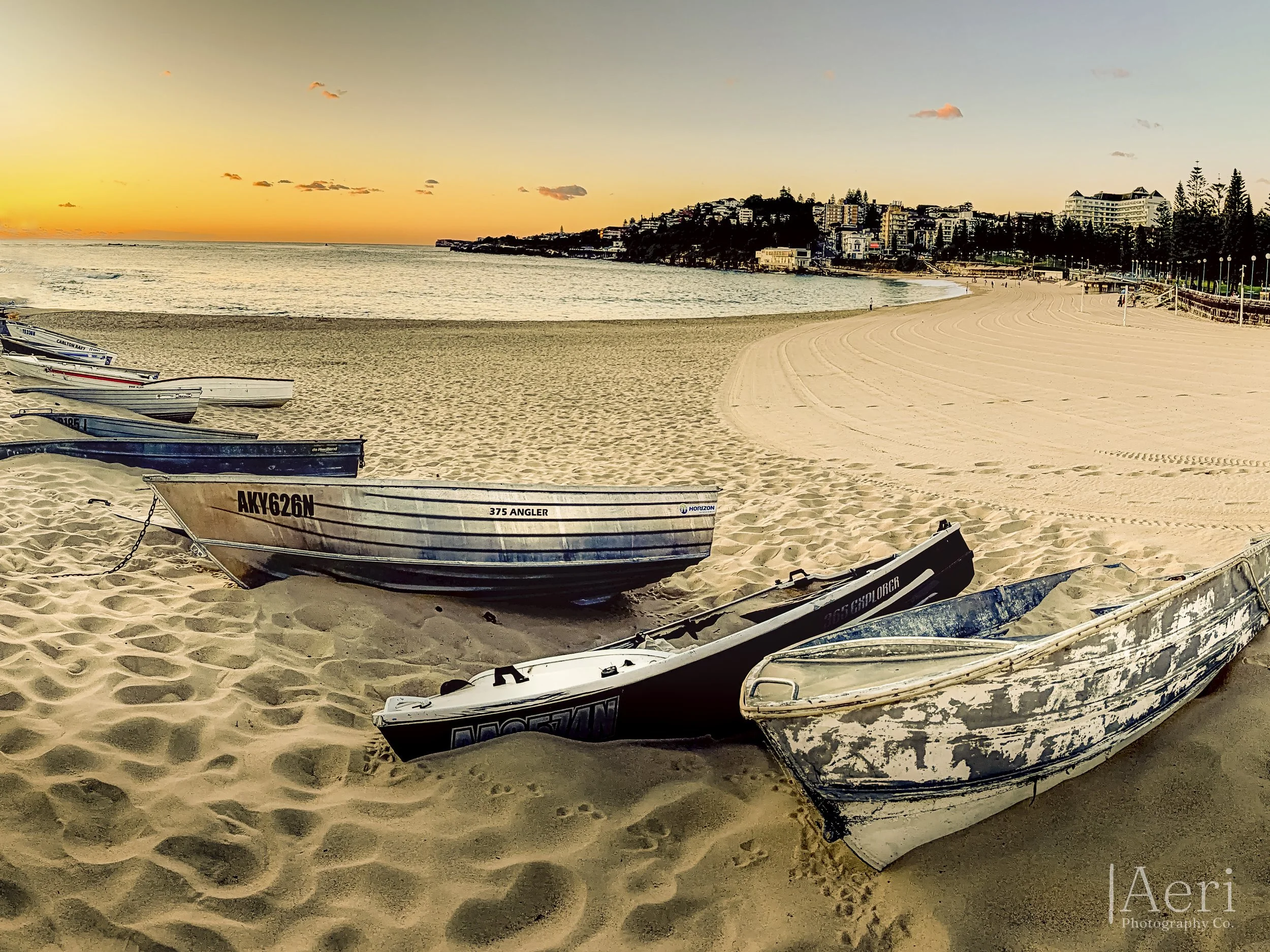 Several little boats are pulled up on the sandy beach at sunset, with a cityscape and calm ocean in the background.