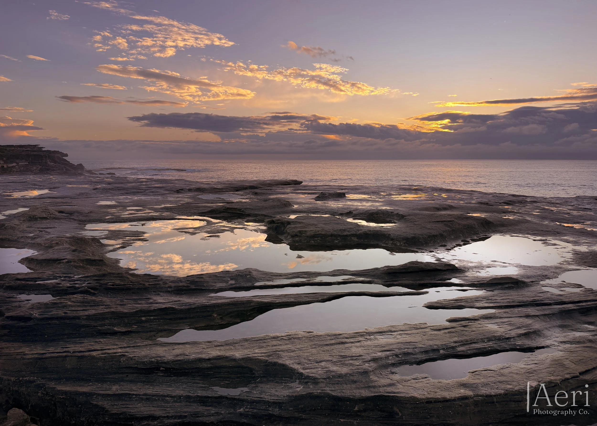 Sunset over a rocky shoreline with tide pools reflecting the sky, clouds, and fading sunlight.