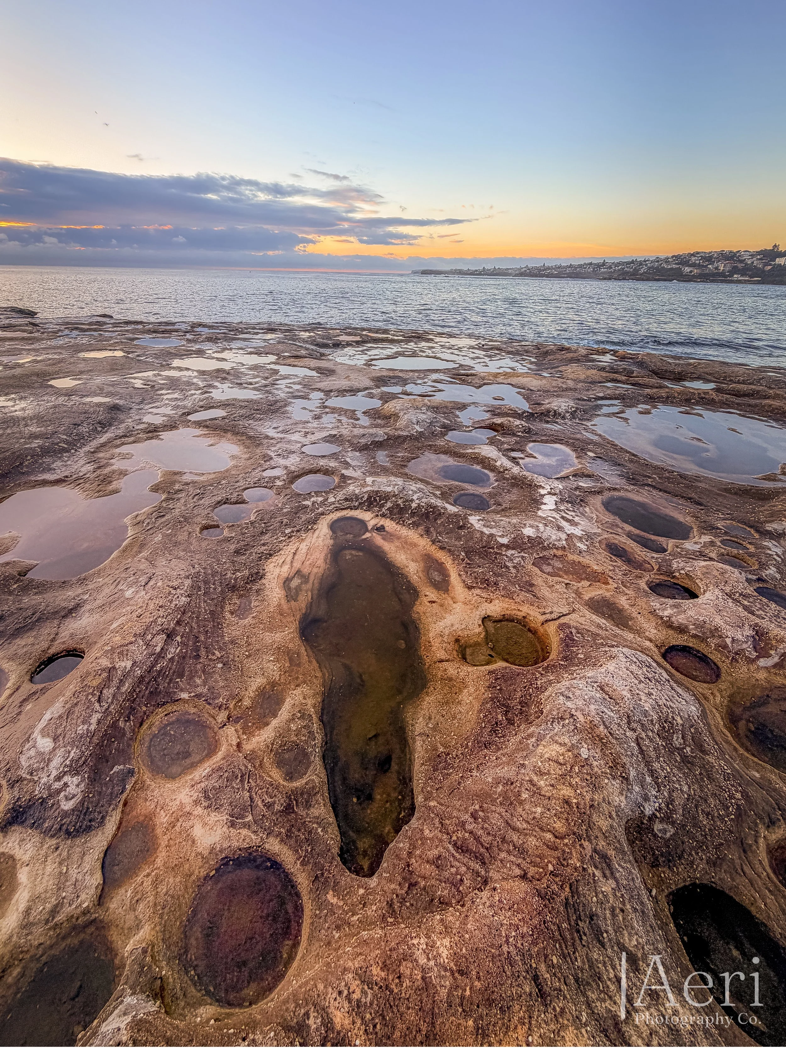 Rocky shoreline with numerous small tide pools during sunset, overlooking the ocean.
