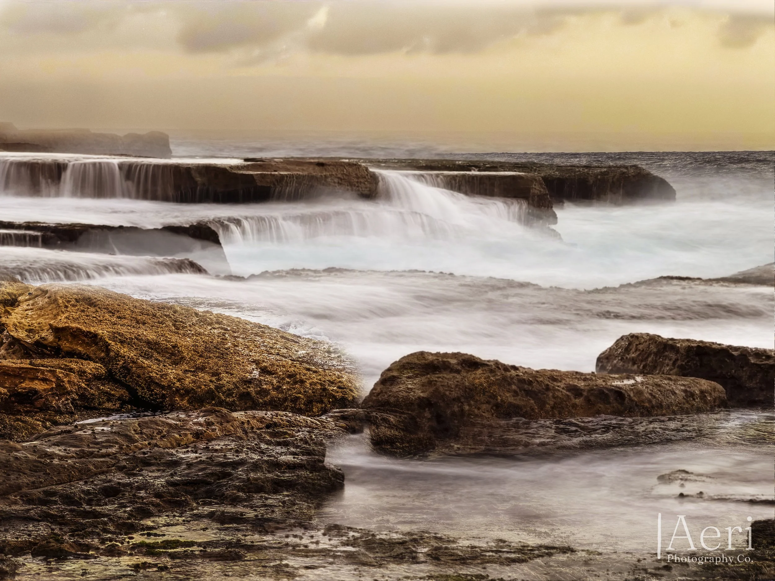 Ocean waves crashing over rocks on a rocky shore during sunset, with a cloudy sky in the background.