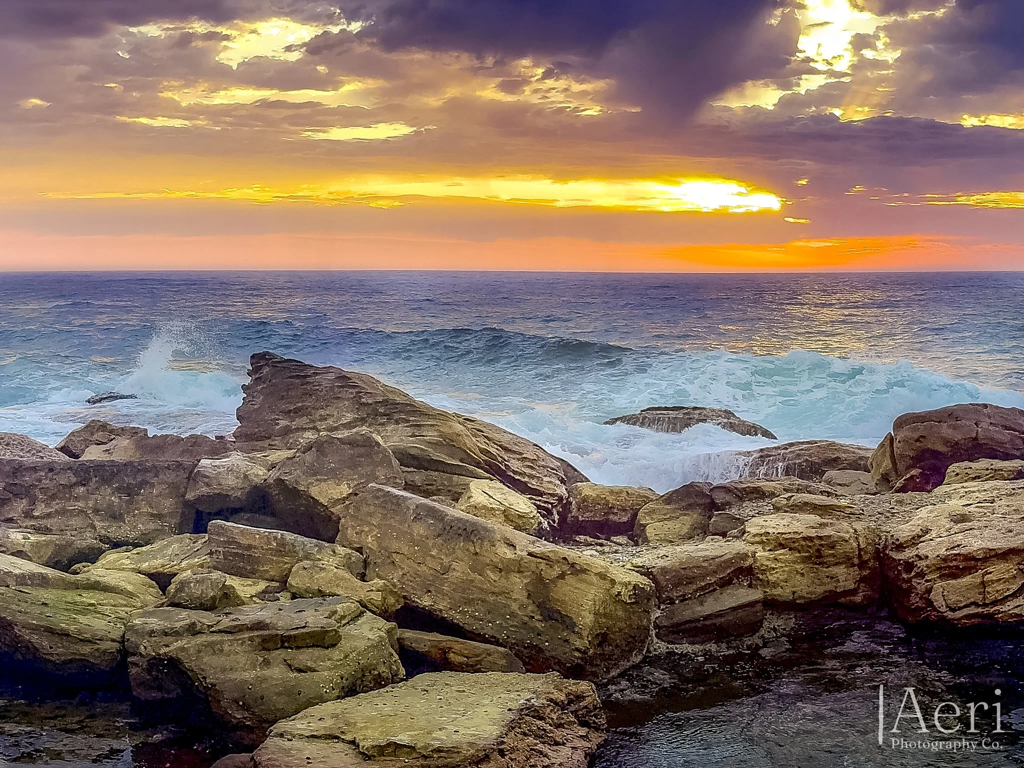 Sunset over the ocean with waves crashing against rocky shoreline.