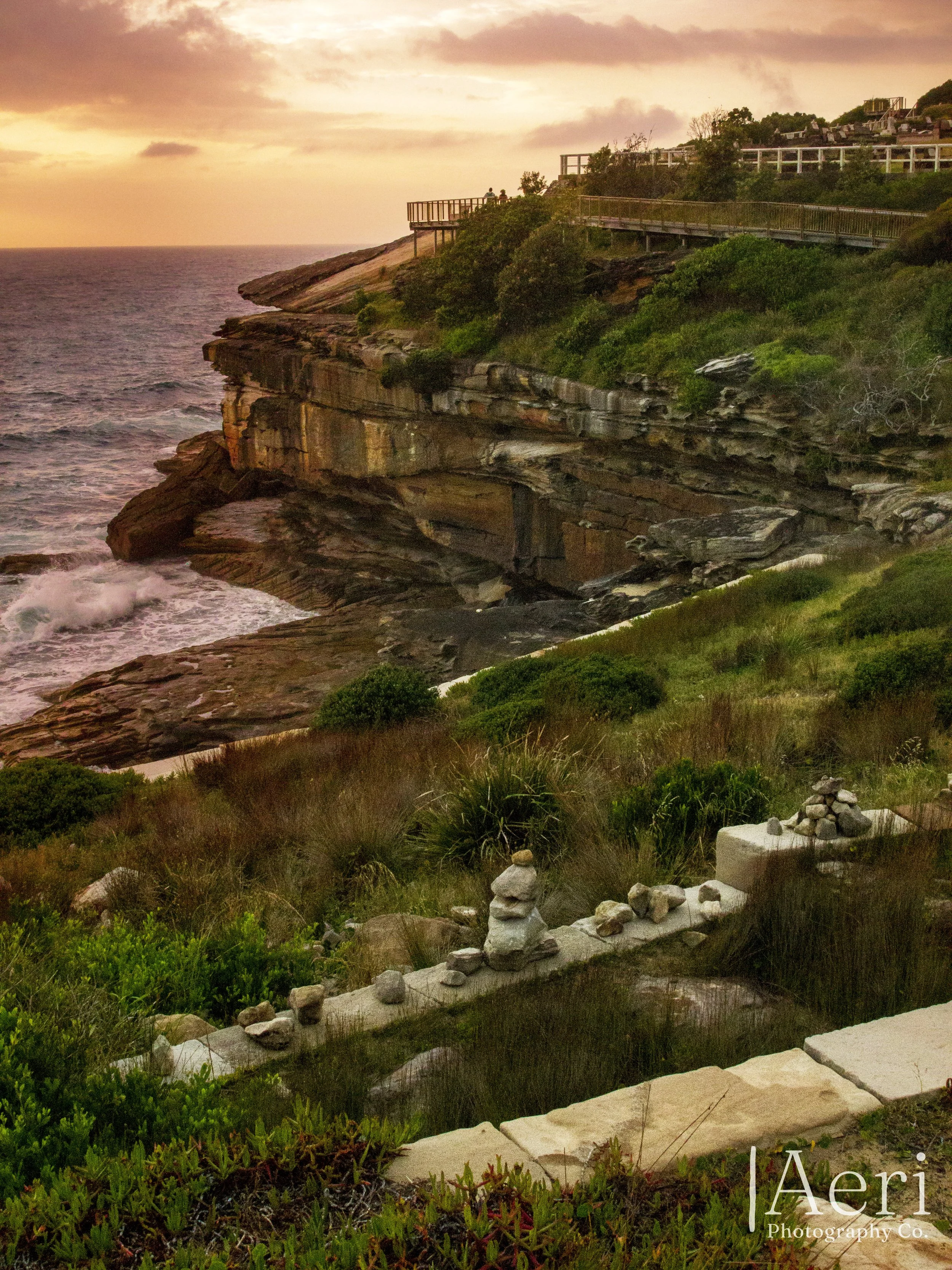 Sunset over coastal cliffs with a walking path, greenery, rock formations, and a view of the ocean.