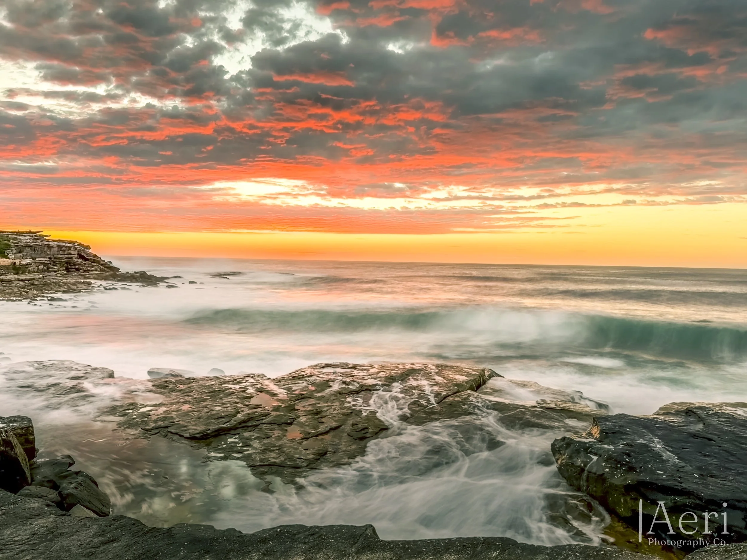 Sunset over the ocean with colorful clouds and waves crashing on rocky shore