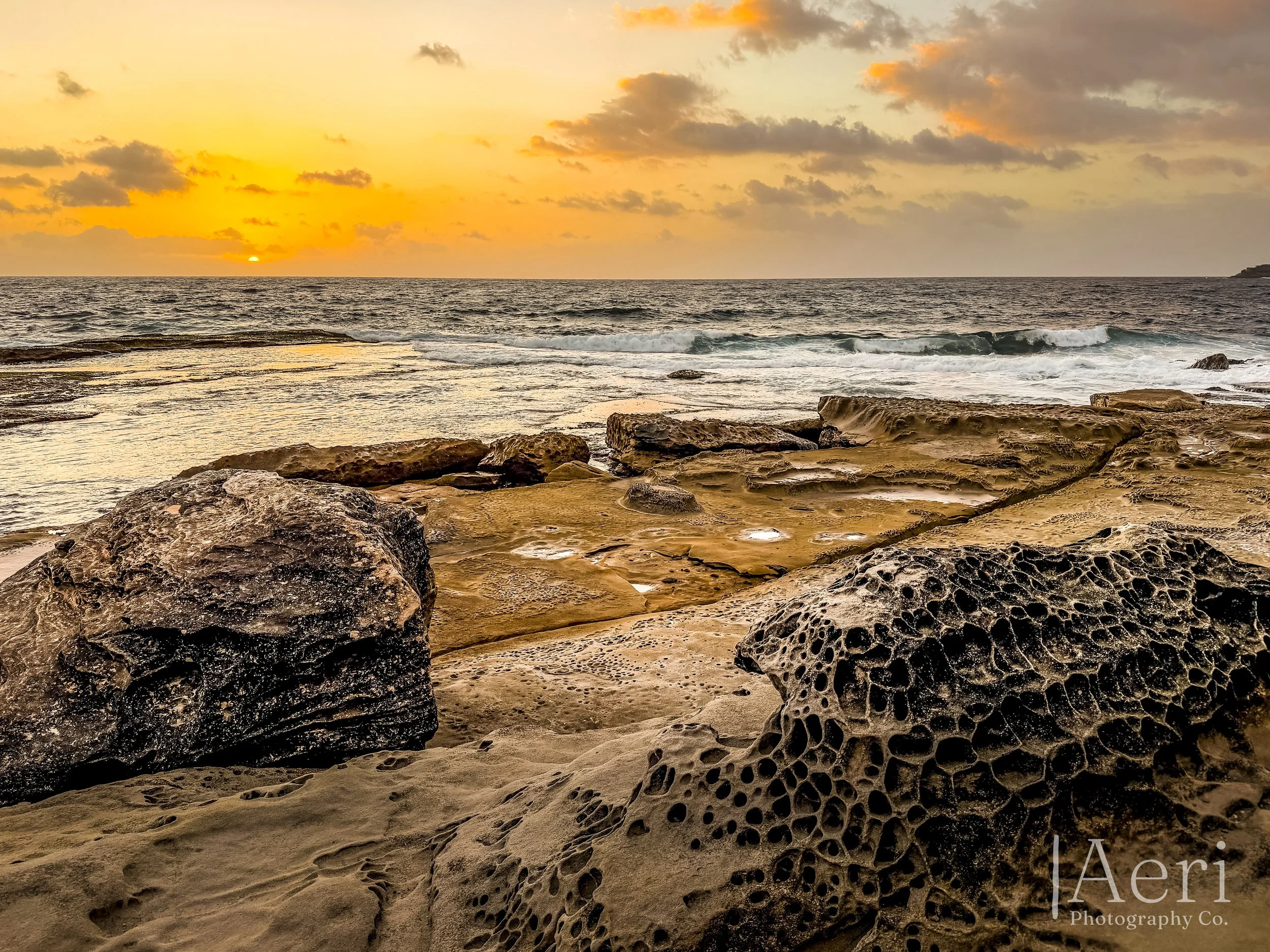 Sunset over a rocky beachfront with large textured rocks and calm ocean waves.