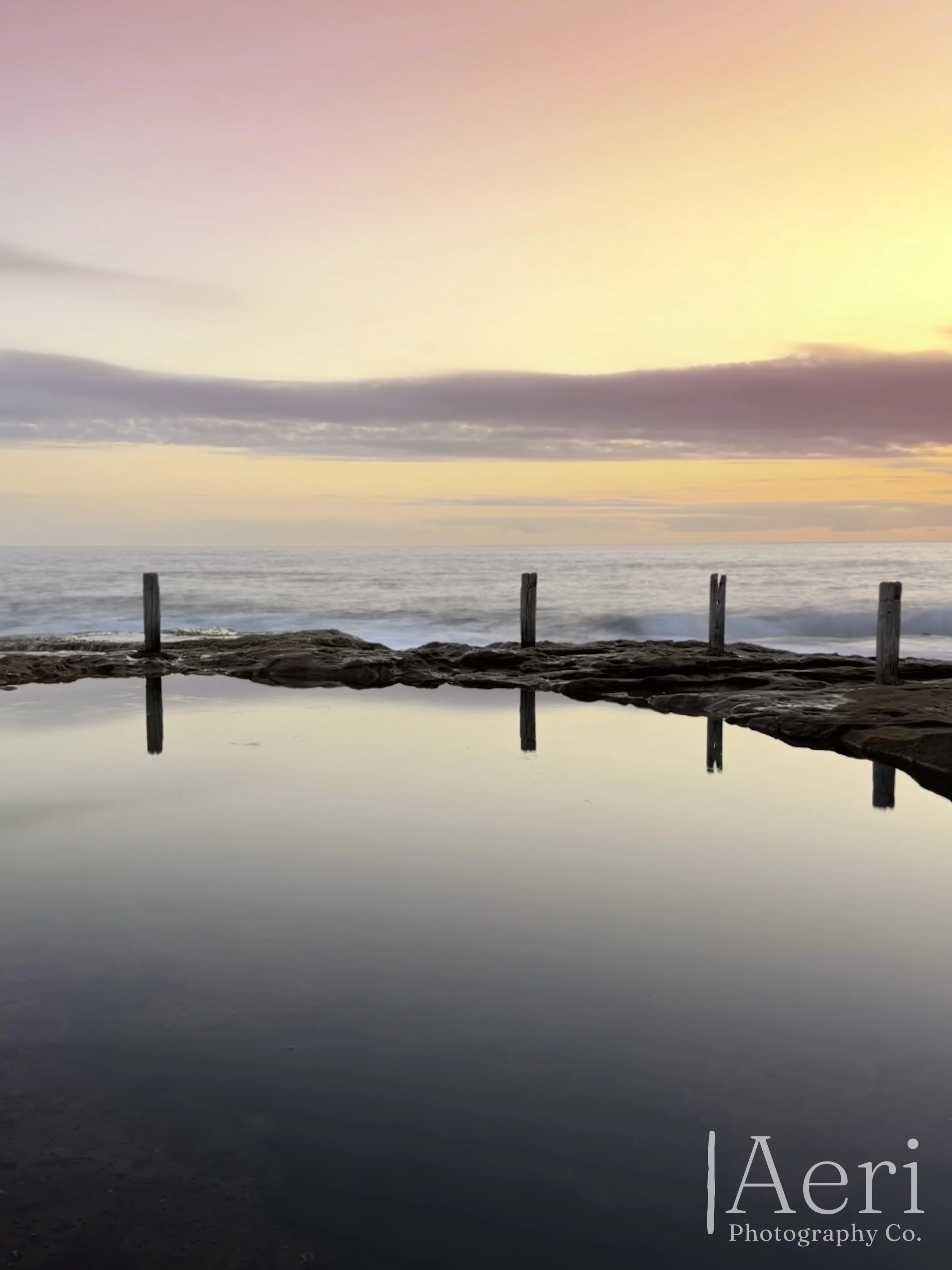 Sunset over the ocean with a rocky foreground and a reflective pool in the foreground. wooden posts line the horizon. Sky has pink, purple, and yellow hues. Aquatic scene with calm water.