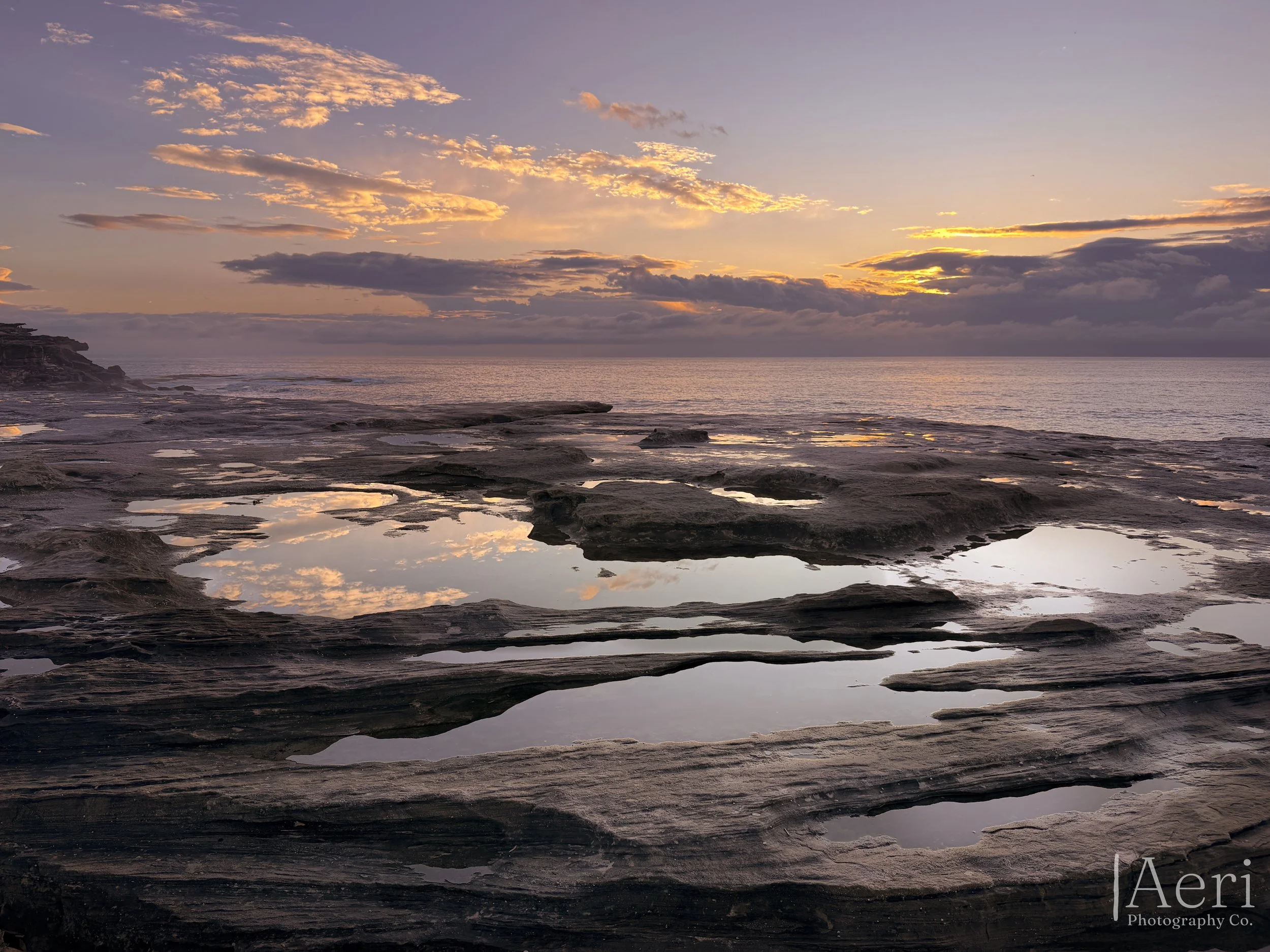 A serene coastal scene during sunset with water reflecting the colorful sky, rocky shoreline, and clouds.