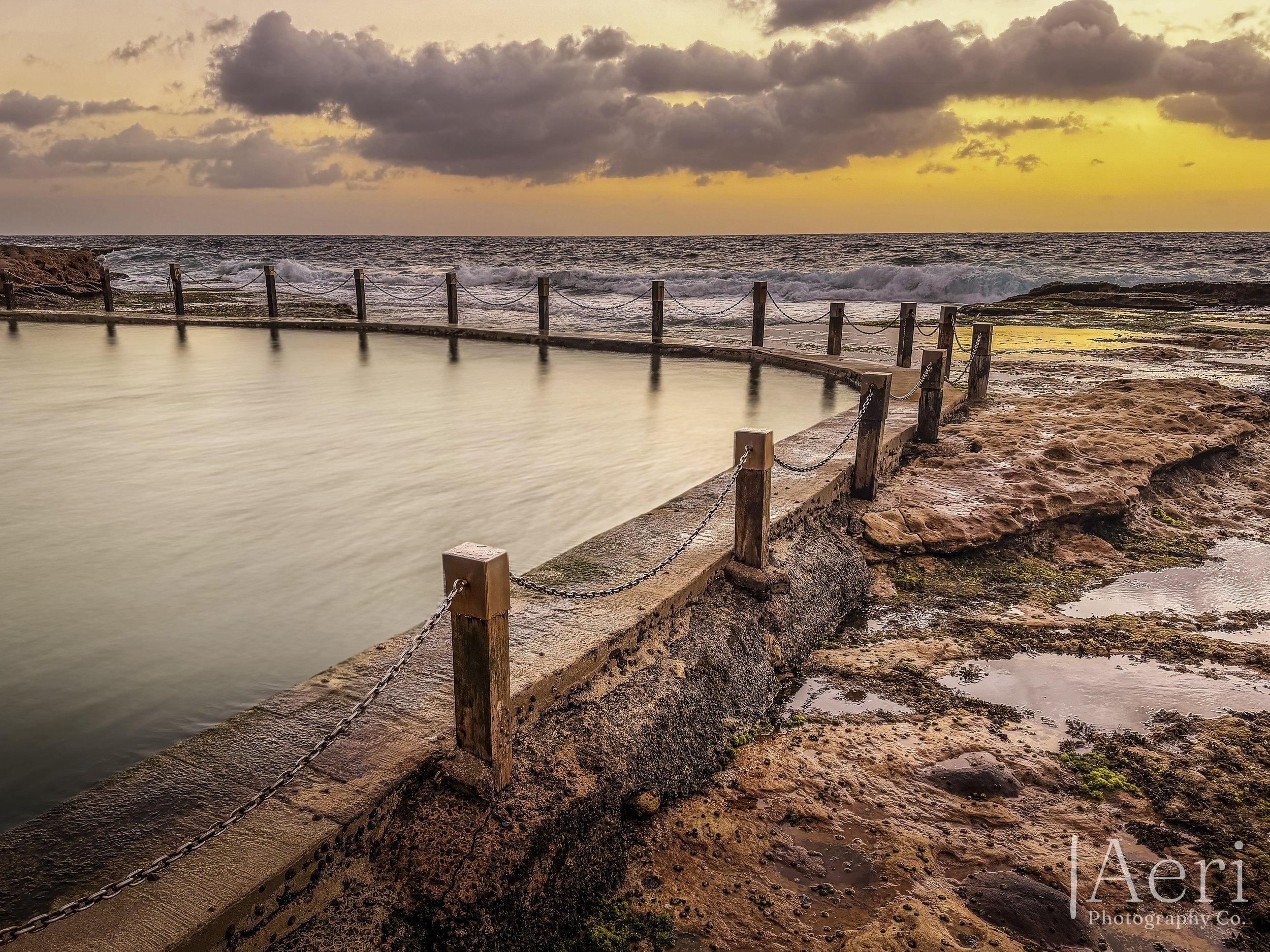 A coastal scene at sunset with a hot spring pool enclosed by a wooden and chain fence, rocky shore, and ocean waves in the background under a cloudy sky.