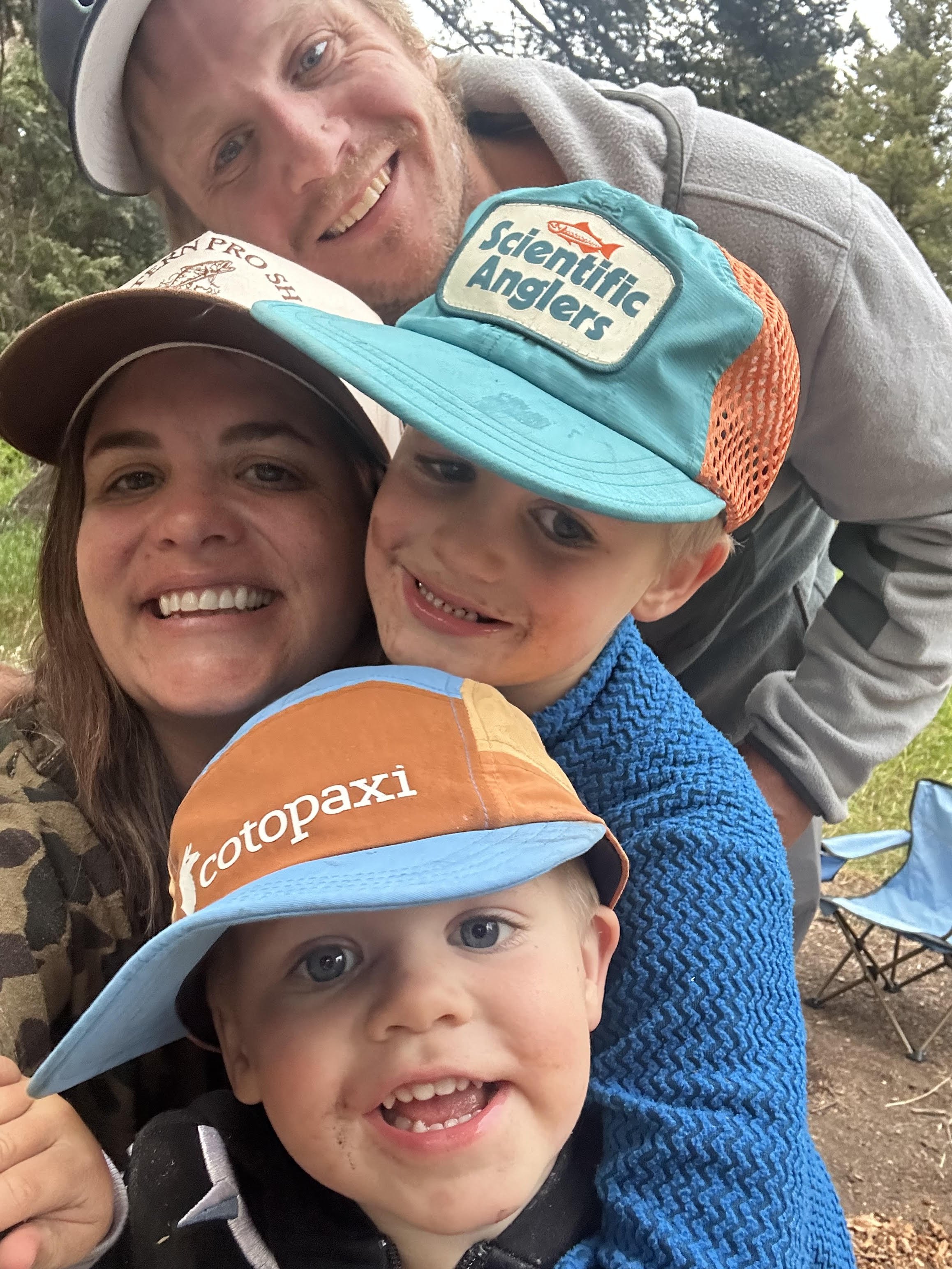 A smiling family of four posed closely together outdoors, wearing hats with outdoor and fishing brand logos. The background shows trees and outdoor camping chairs.
