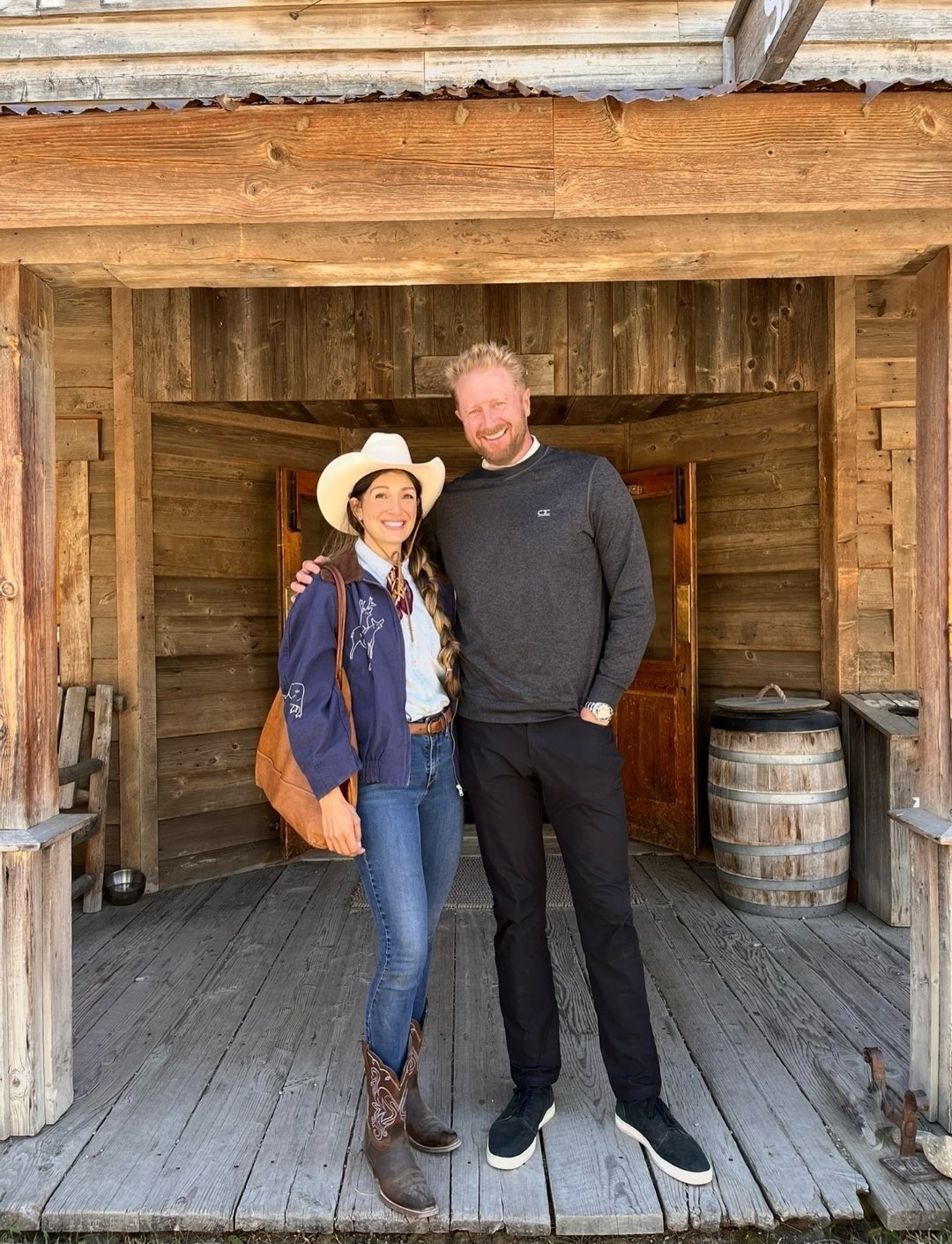 A woman wearing cowboy boots, a white cowboy hat, and a blue jacket stands with a man in a black sweatshirt, both smiling, on a wooden porch with rustic wooden walls and a barrel in the background.