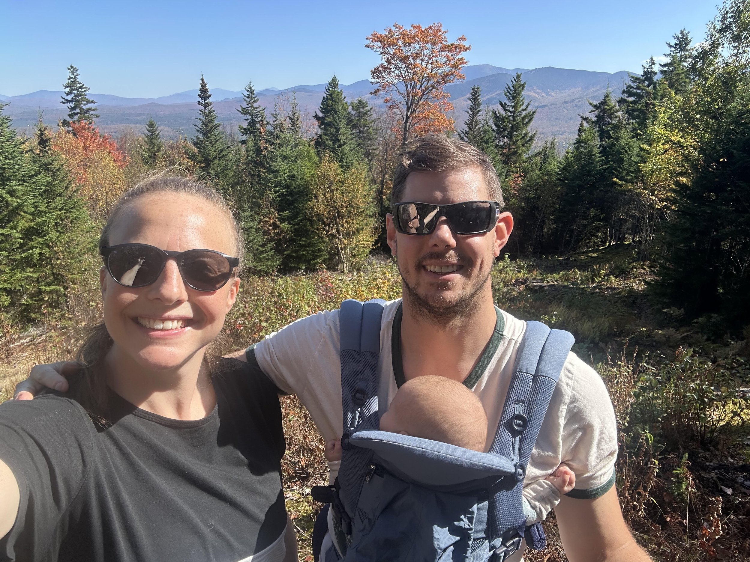 A smiling woman and a man wearing sunglasses are taking a selfie outdoors in a forested area with mountains in the background. The man is carrying a baby in a front-facing carrier.