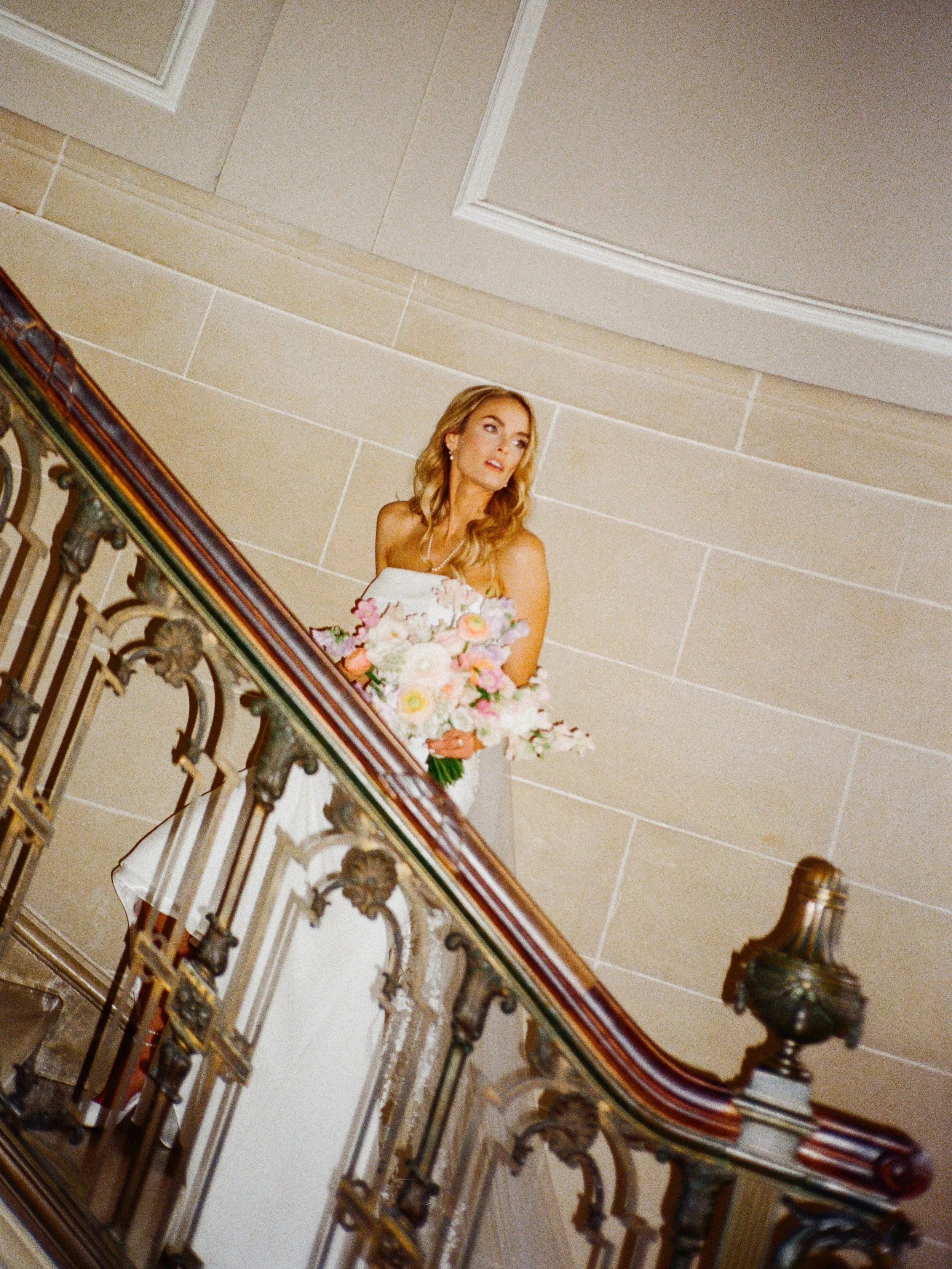 A woman holding a bouquet of flowers, standing on a staircase with an ornate wooden and metal railing, looking to the side in an elegant indoor setting.