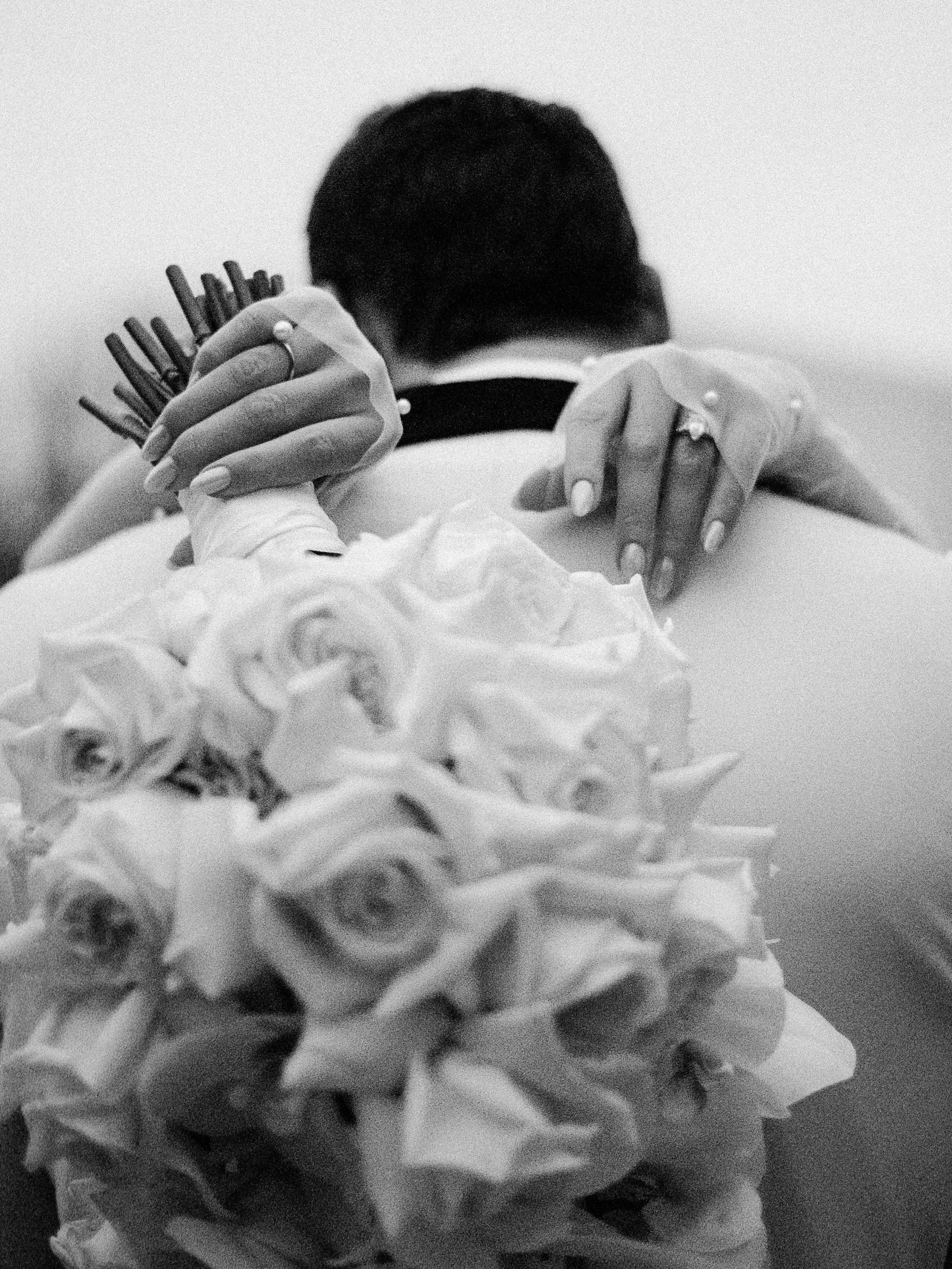 A person with short hair hugging another person. The person being hugged has hands with rings and manicured nails resting on the other's shoulder. A bouquet of roses is in the foreground.