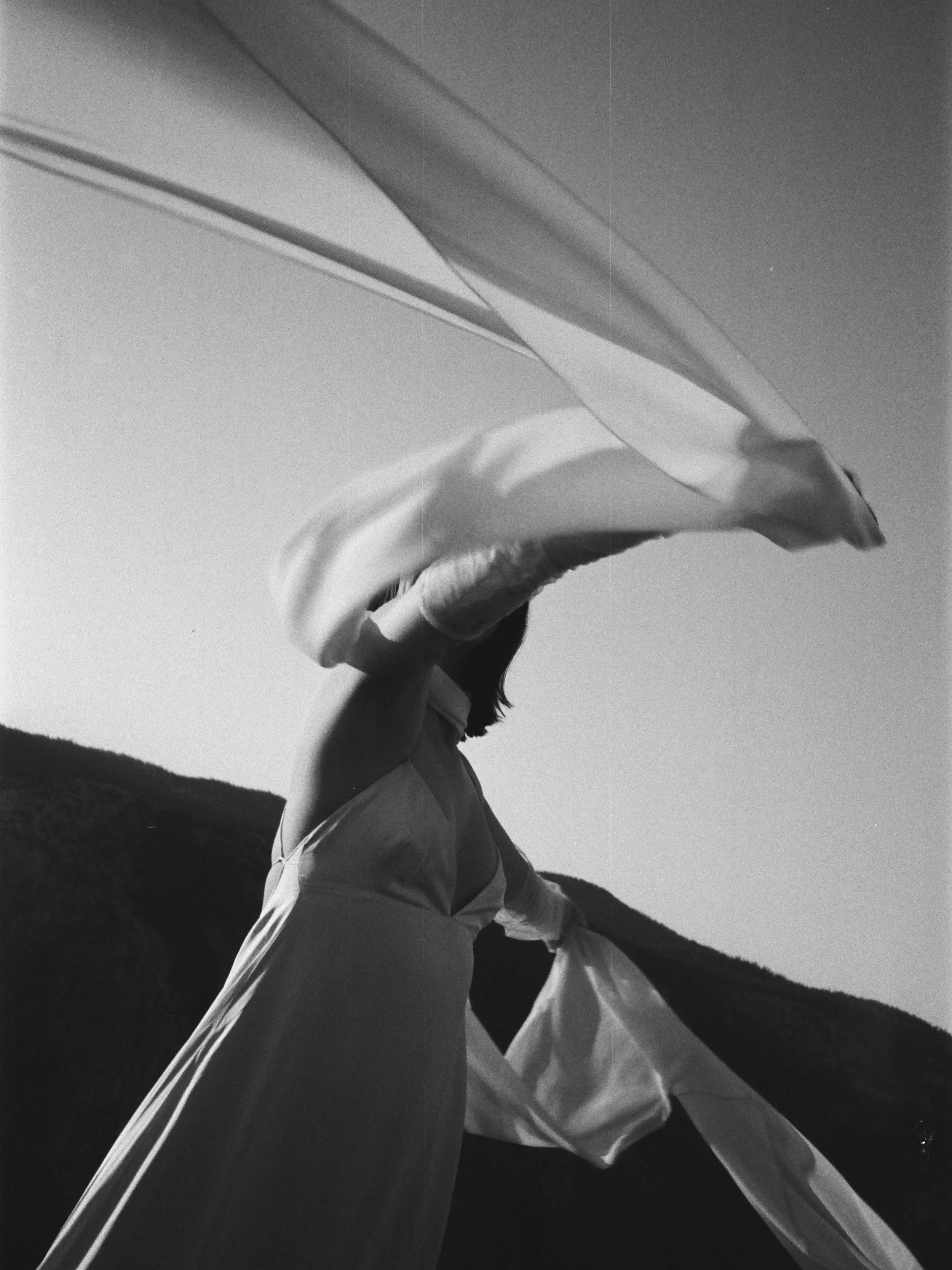 Woman holding flowing fabric outdoors with a tree-covered hill in the background, black and white photo