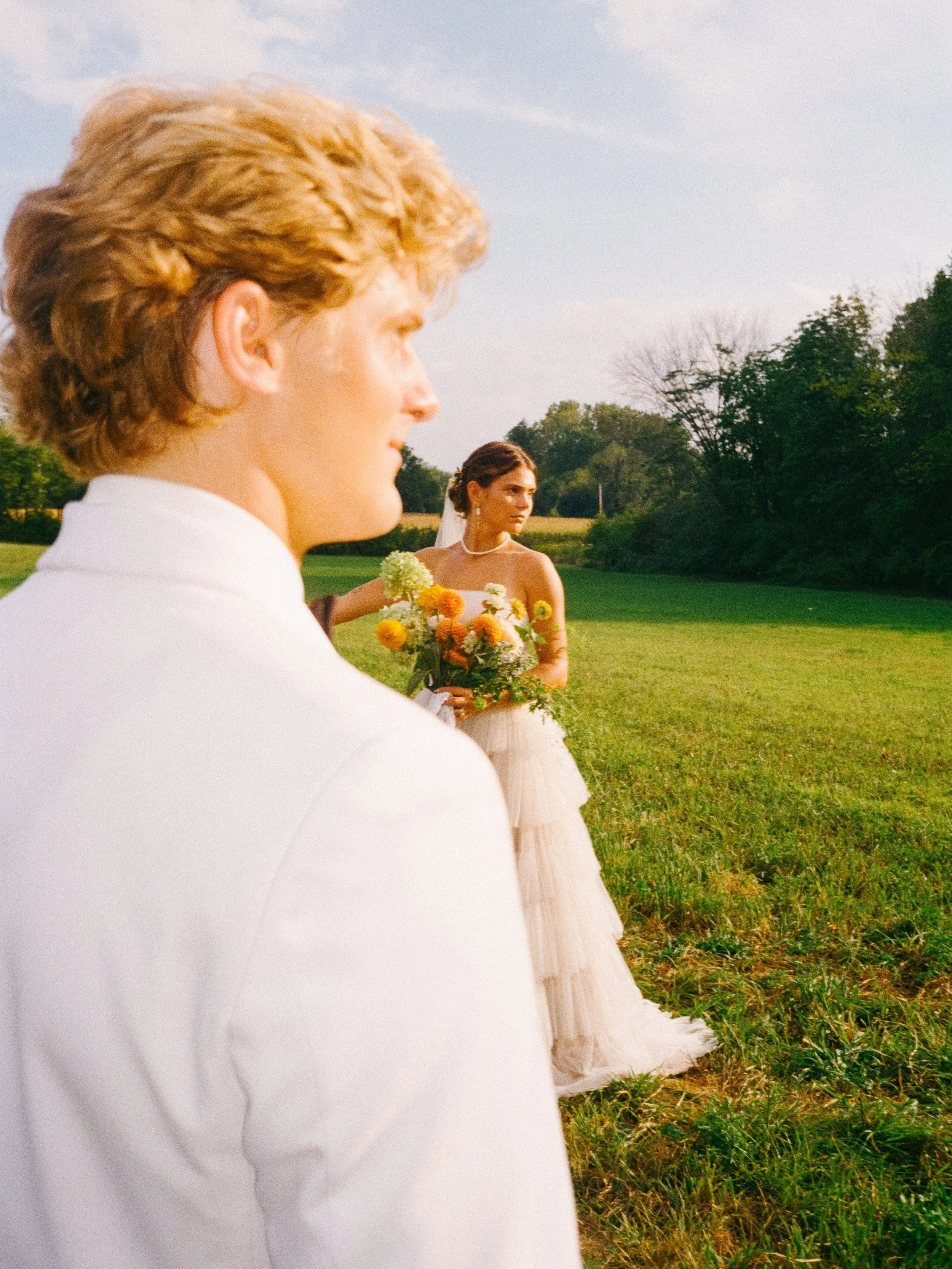 A groom with blond hair in a white suit in the foreground, and a bride with dark hair and a strapless wedding dress holding a bouquet of yellow and orange flowers, standing on a grassy field with trees in the background on a sunny day.