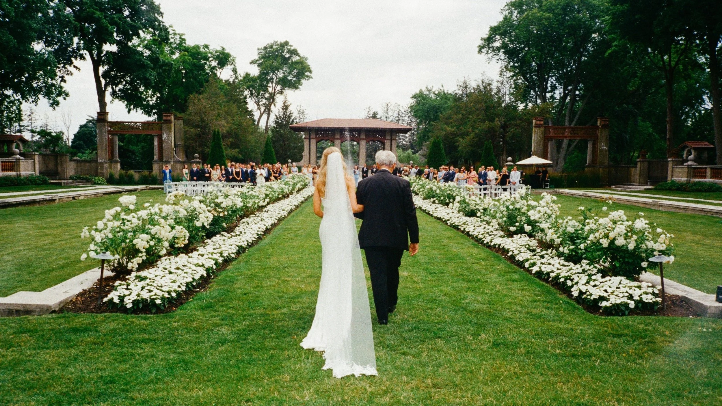 A bride in a white wedding dress walking down a grassy aisle with an older man, with guests gathered in the background in a lush green outdoor setting.
