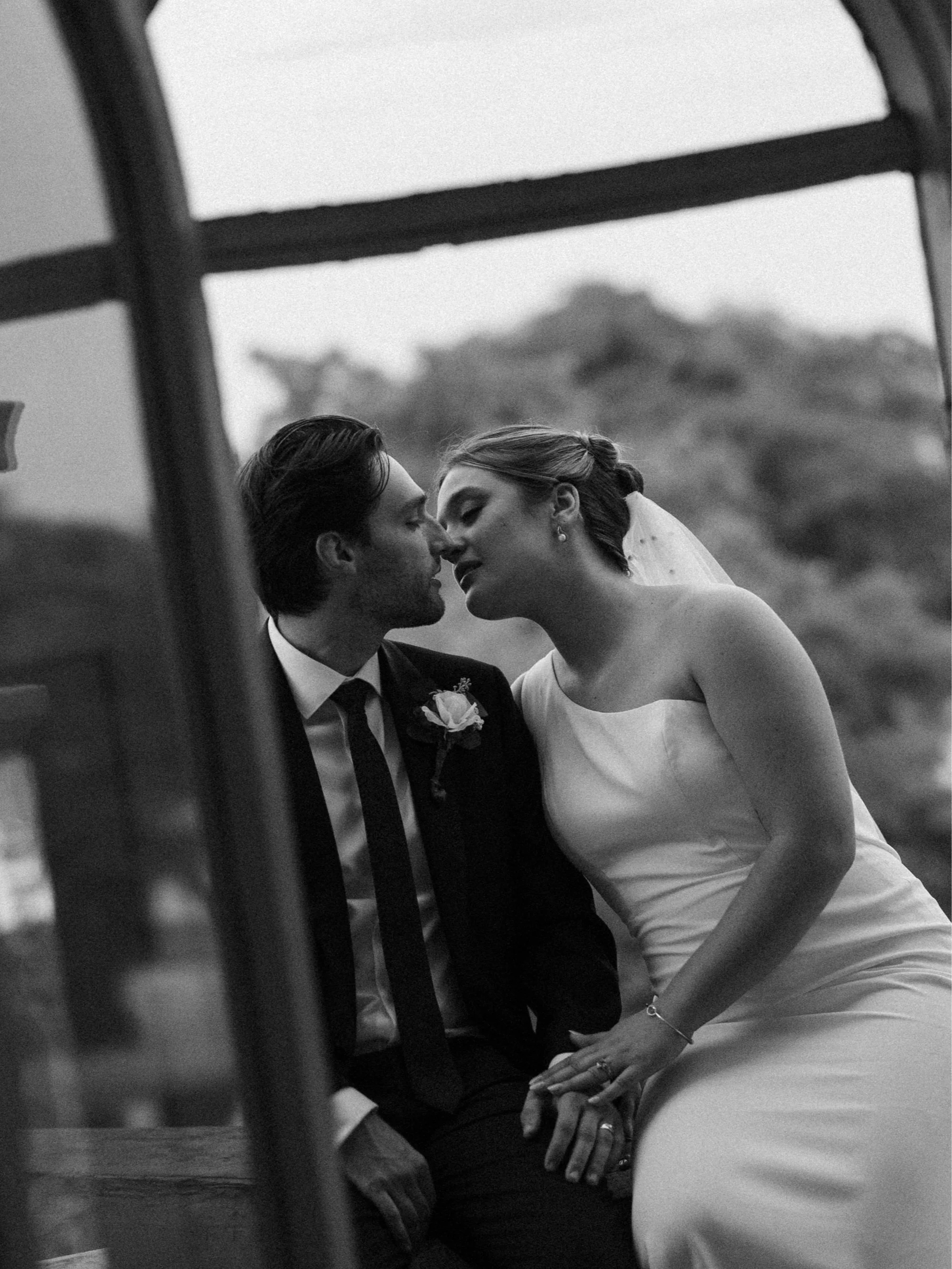 Black and white photo of a bride and groom close to each other, leaning in for a kiss, sitting on a bench with a blurred outdoor landscape in the background.