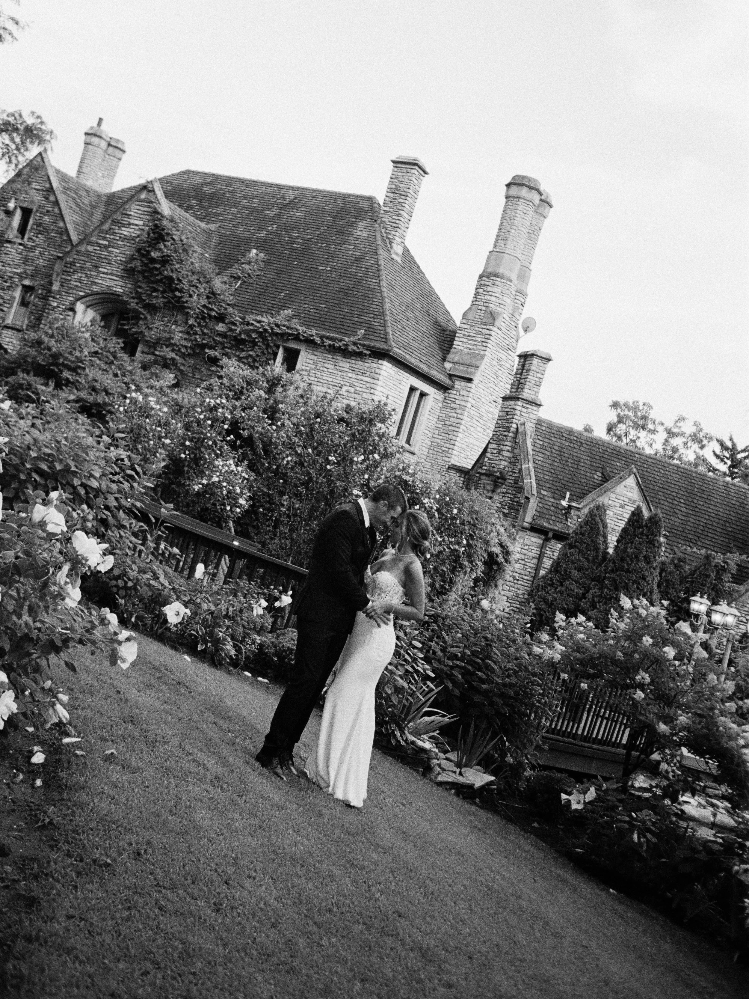 A bride and groom in wedding attire standing close together in a garden with a large, historic brick house with multiple chimneys in the background.