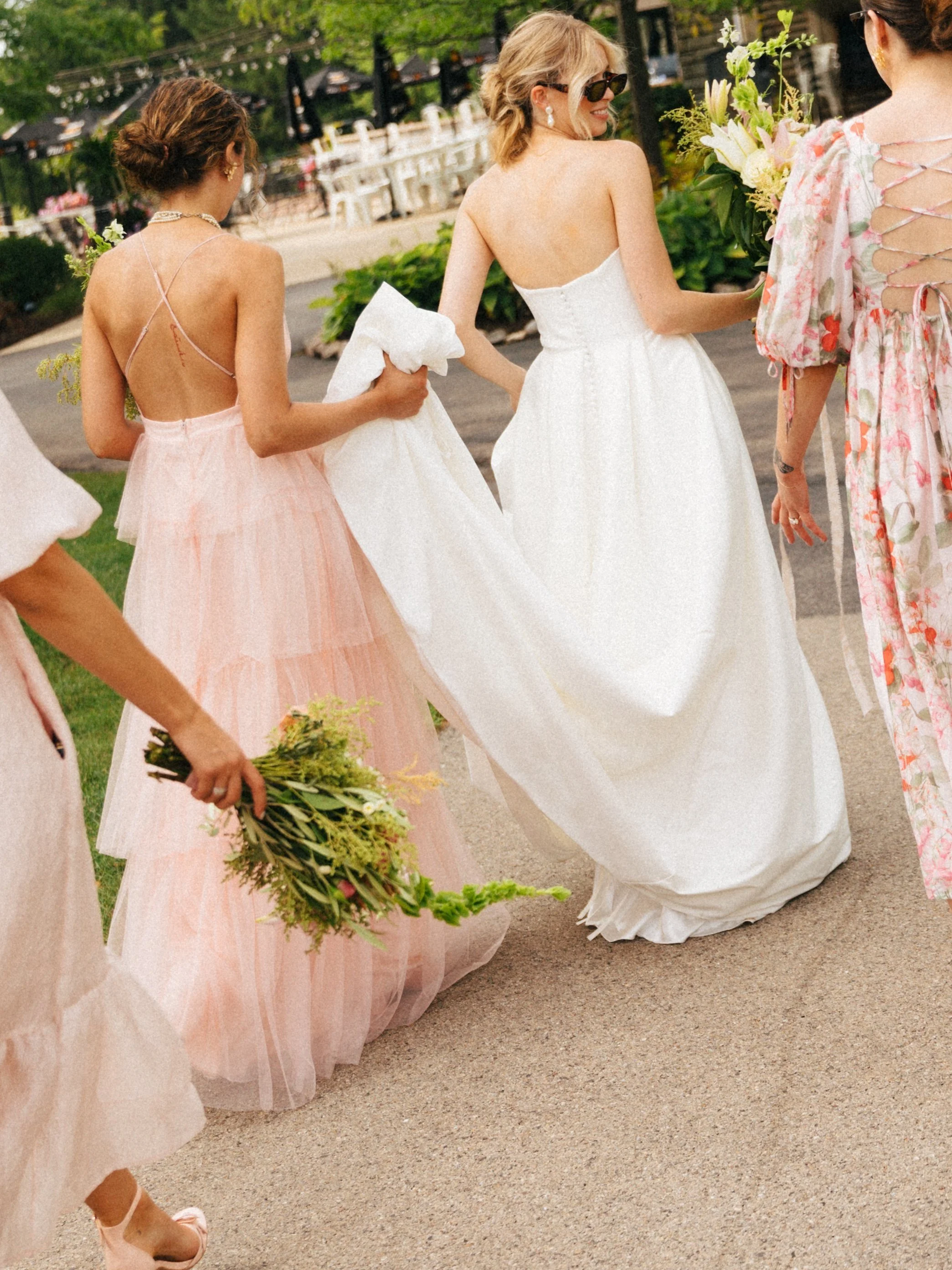 Group of women, one in a white wedding dress, at a wedding outdoor event, holding bouquets of flowers, with some women wearing pink dresses and one in a floral dress.