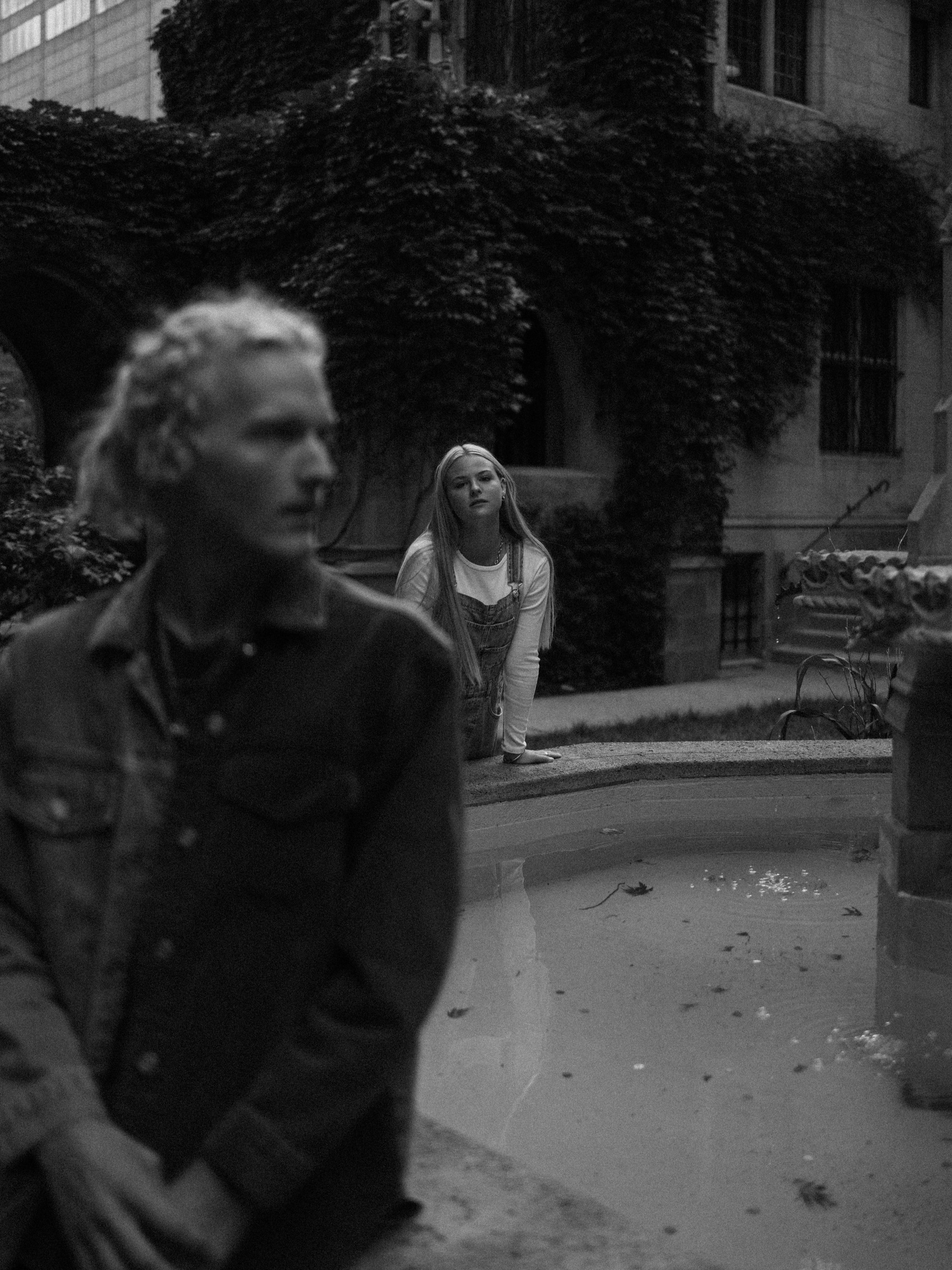 A young girl with long hair looking at a woman in the foreground near a fountain in an outdoor courtyard.