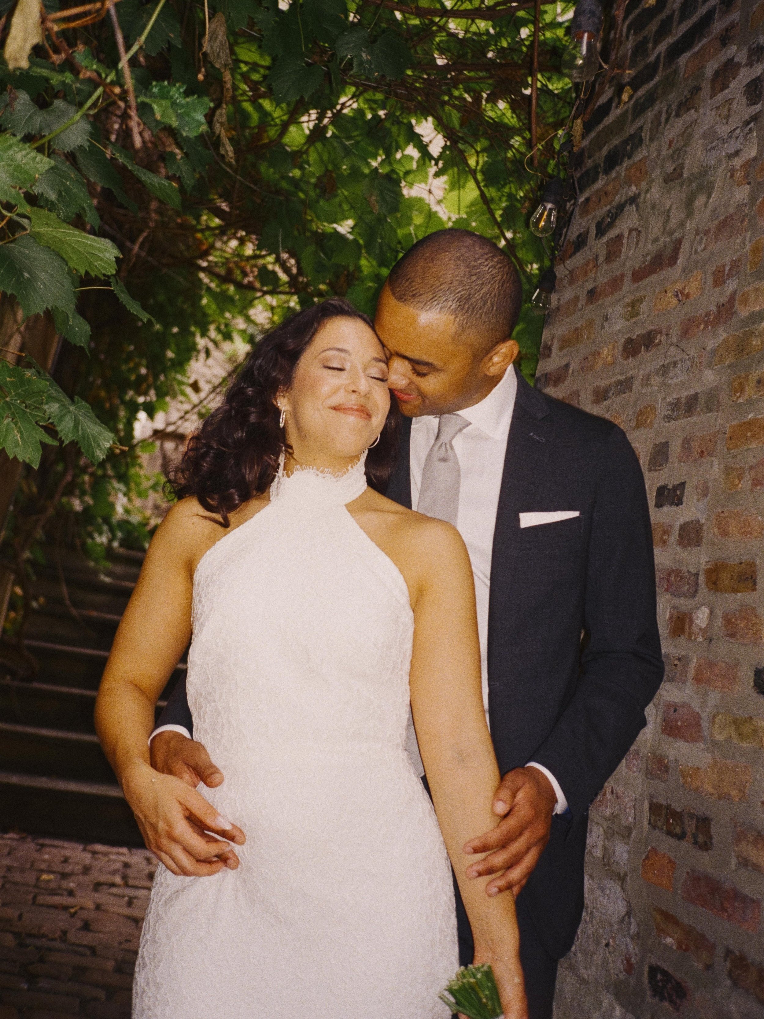 A bride and groom share a tender moment, standing close with the groom gently holding the bride's waist and the bride smiling contentedly with eyes closed. They are outside, next to a brick wall and surrounded by green foliage.