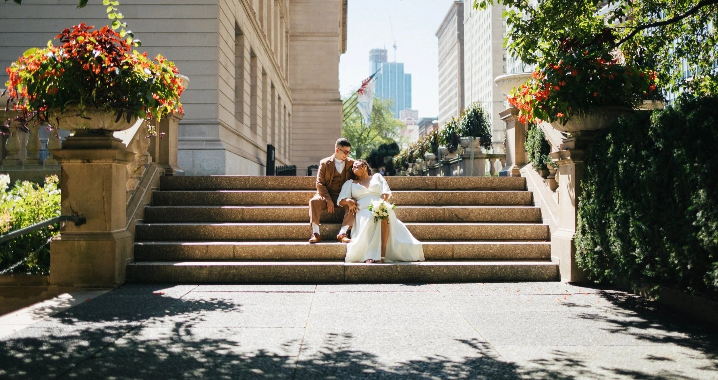 A couple in wedding attire sitting on outdoor steps surrounded by large flower arrangements and city buildings in the background.