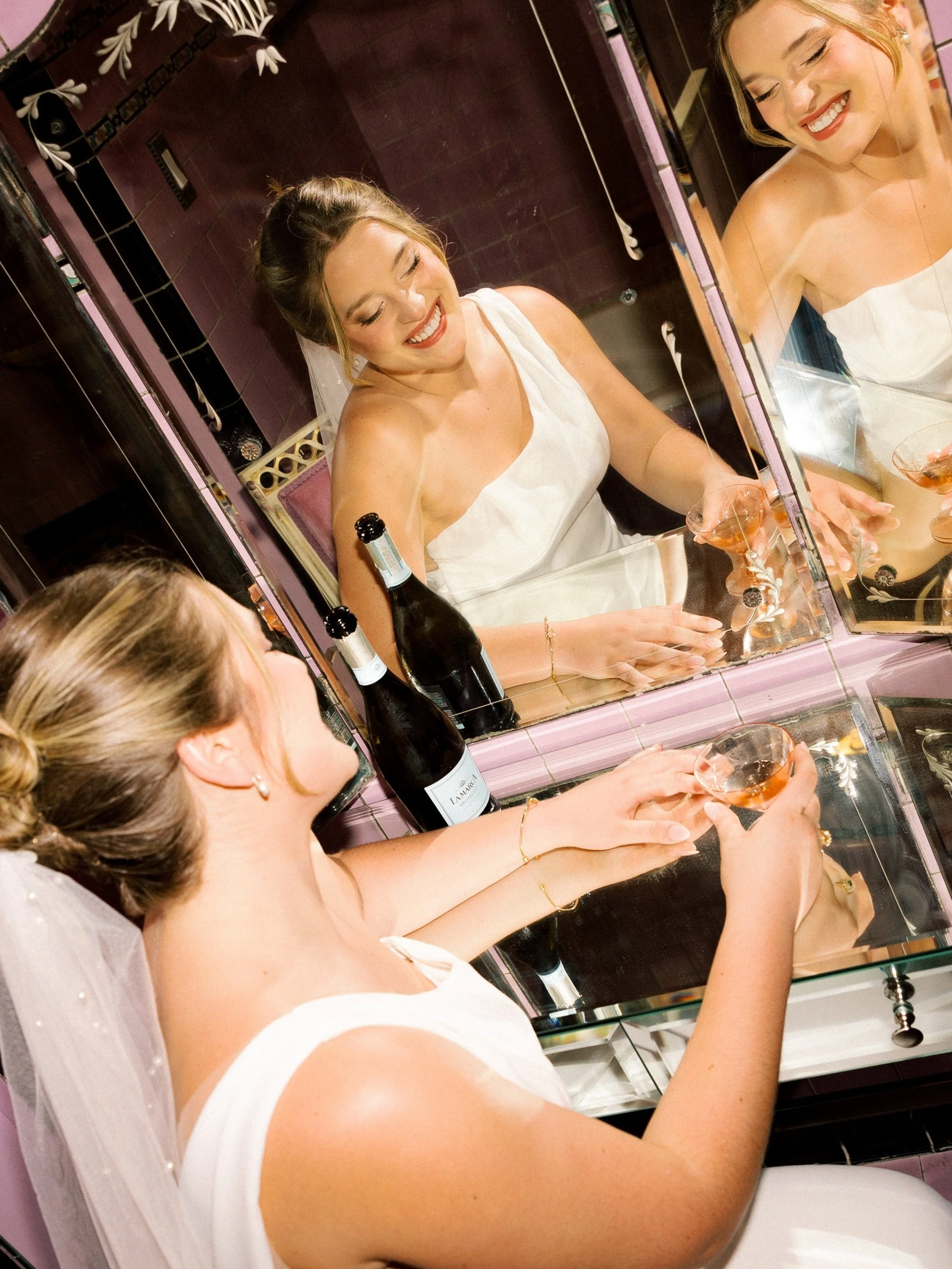 A bride with a veil and wedding jewelry looking at herself in a mirror, holding a glass of rosé wine, with champagne bottles on the table in front of her.