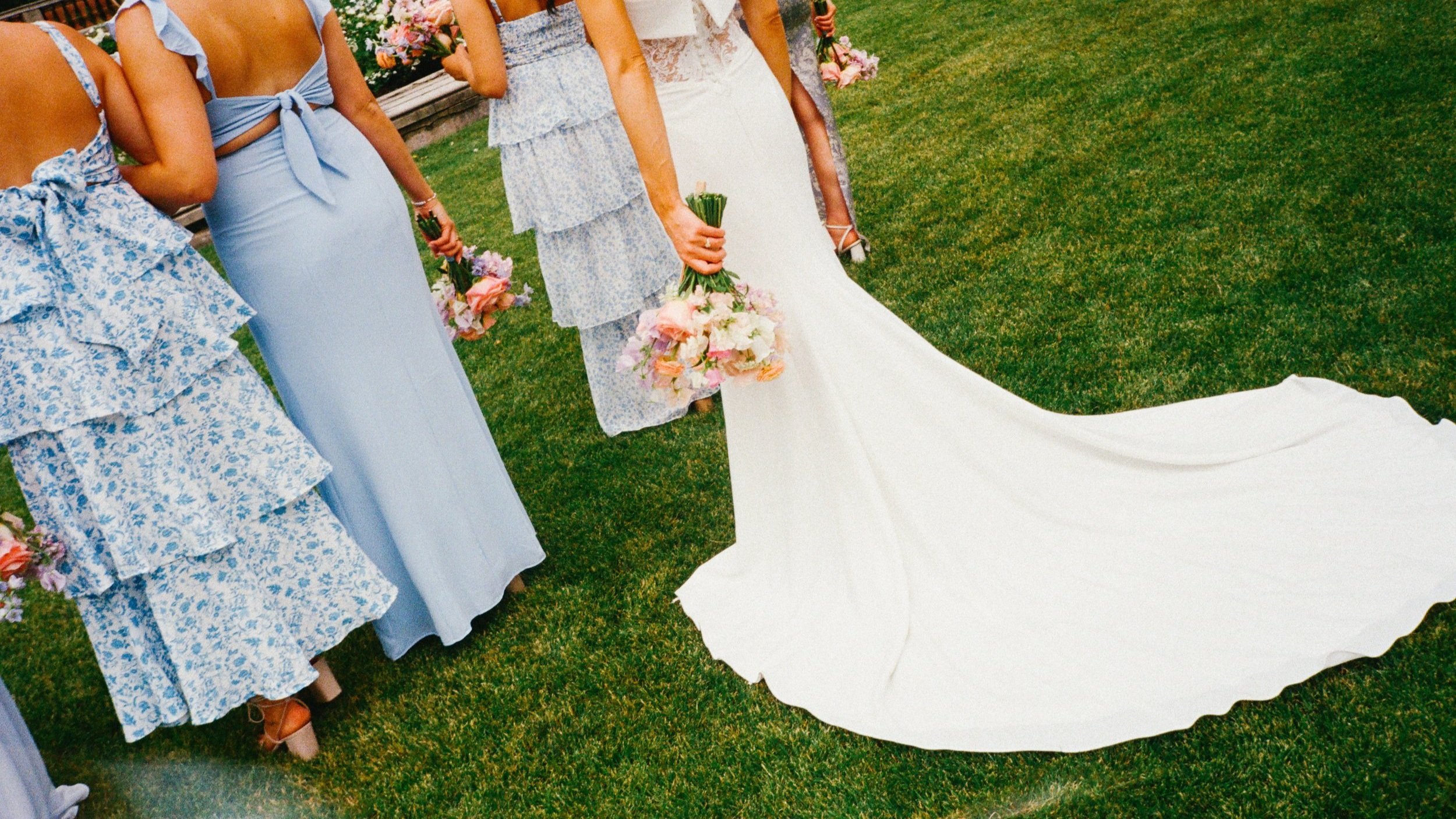 A bride in a white wedding gown holding a bouquet of pink and white flowers, standing on green grass during a wedding ceremony with bridesmaids dressed in blue and floral dresses holding similar bouquets.