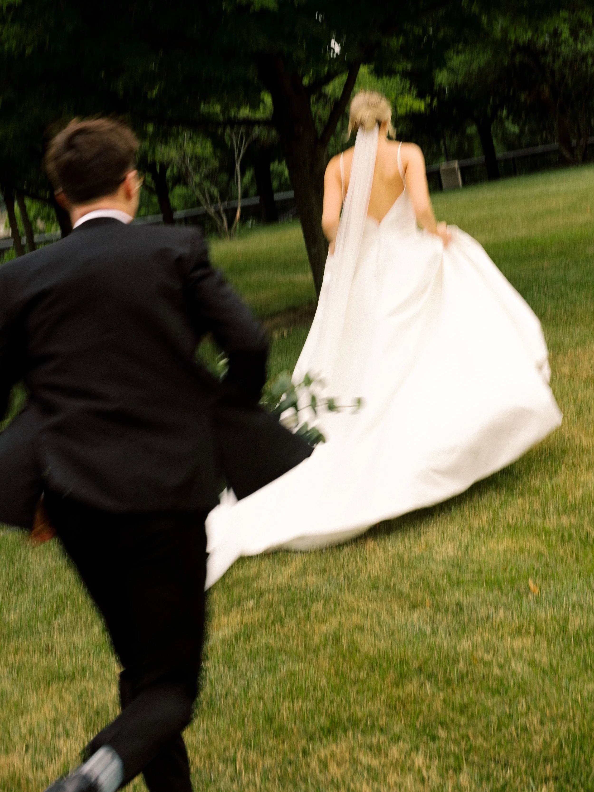 Blurry photo of a bride in a white wedding dress walking away, with a man in a black suit running towards her in a grassy outdoor area with trees.