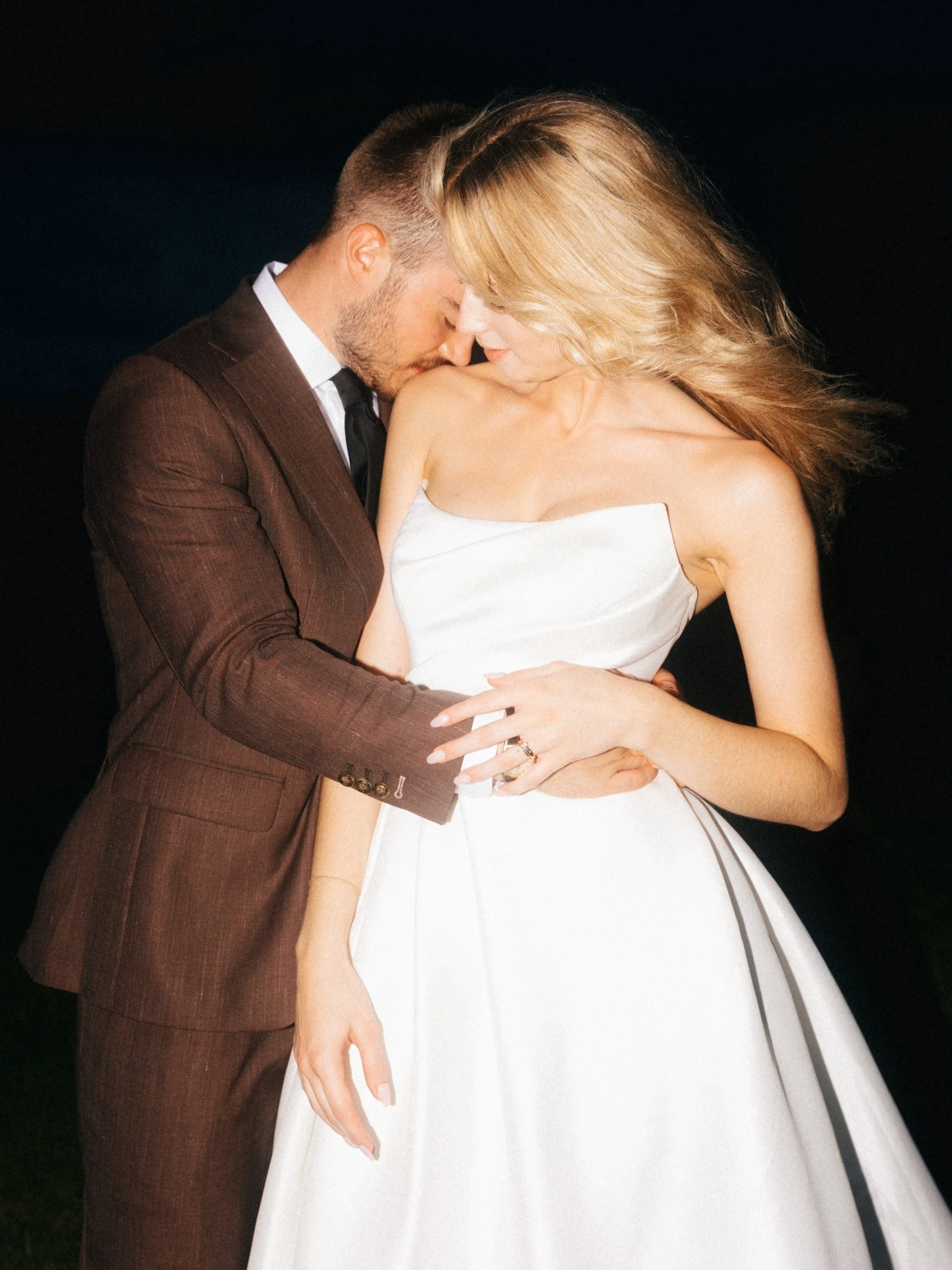 A romantic couple, a man in a brown suit and a woman in a white dress, holding each other closely with their faces touching, against a dark background.